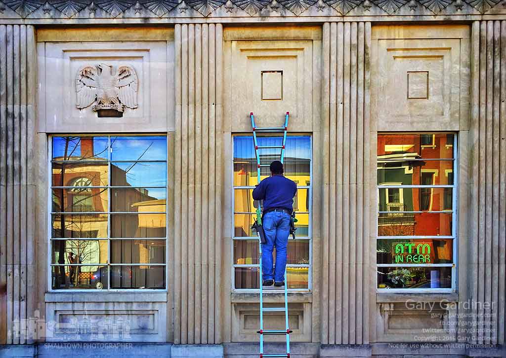 A window washer steadies himself on the ladder while washing the center window of the Middlefield Bank in Uptown Westerville as stores across the street lit by late afternoon sun are reflected in the cleared glass. My Final Photo for Nov. 16, 2015.