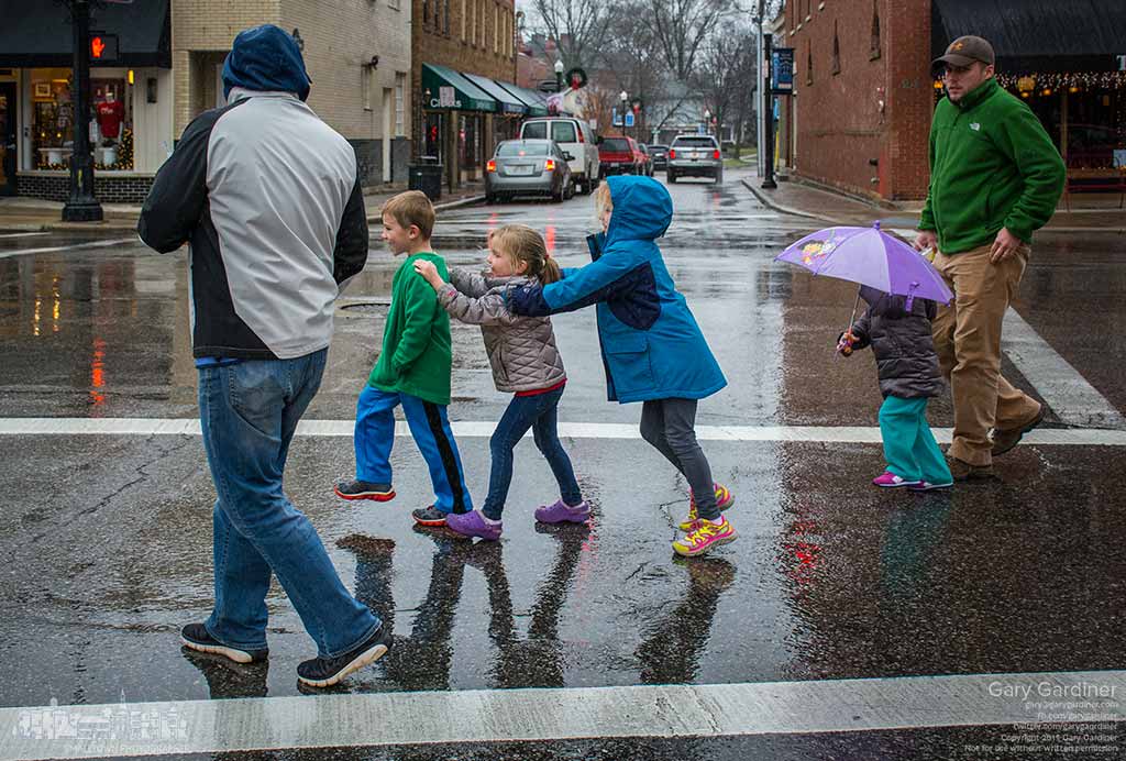 A trio of youngsters holding each other as if they are a train are followed by another under an umbrella as they cross Main Street during a rain storm n Uptown. My Final Photo for Dec. 28, 2015.