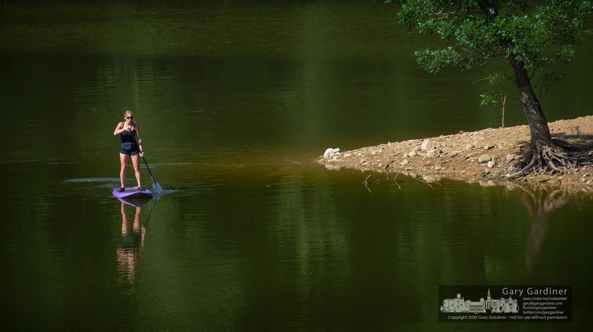 A paddleboarder rounds a small point of land on her way to the Red Bank Road bridge where the boat landing awaits. My Final Photo for July 26, 2020.