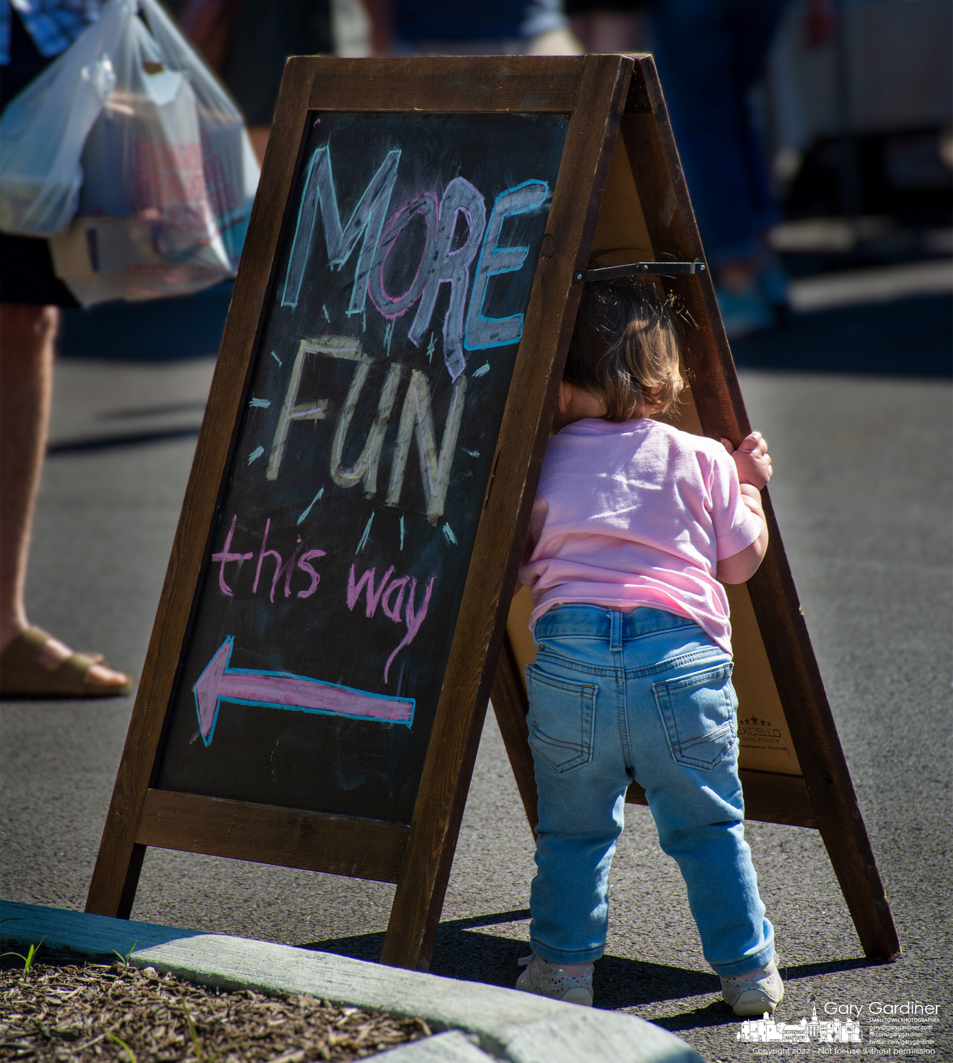 A toddler passes through a signboard indicating a more fun experience than staying with her parents at the Saturday Farmers' Market in Uptown Westerville Saturday morning. My Final Photo for June 18, 2022.