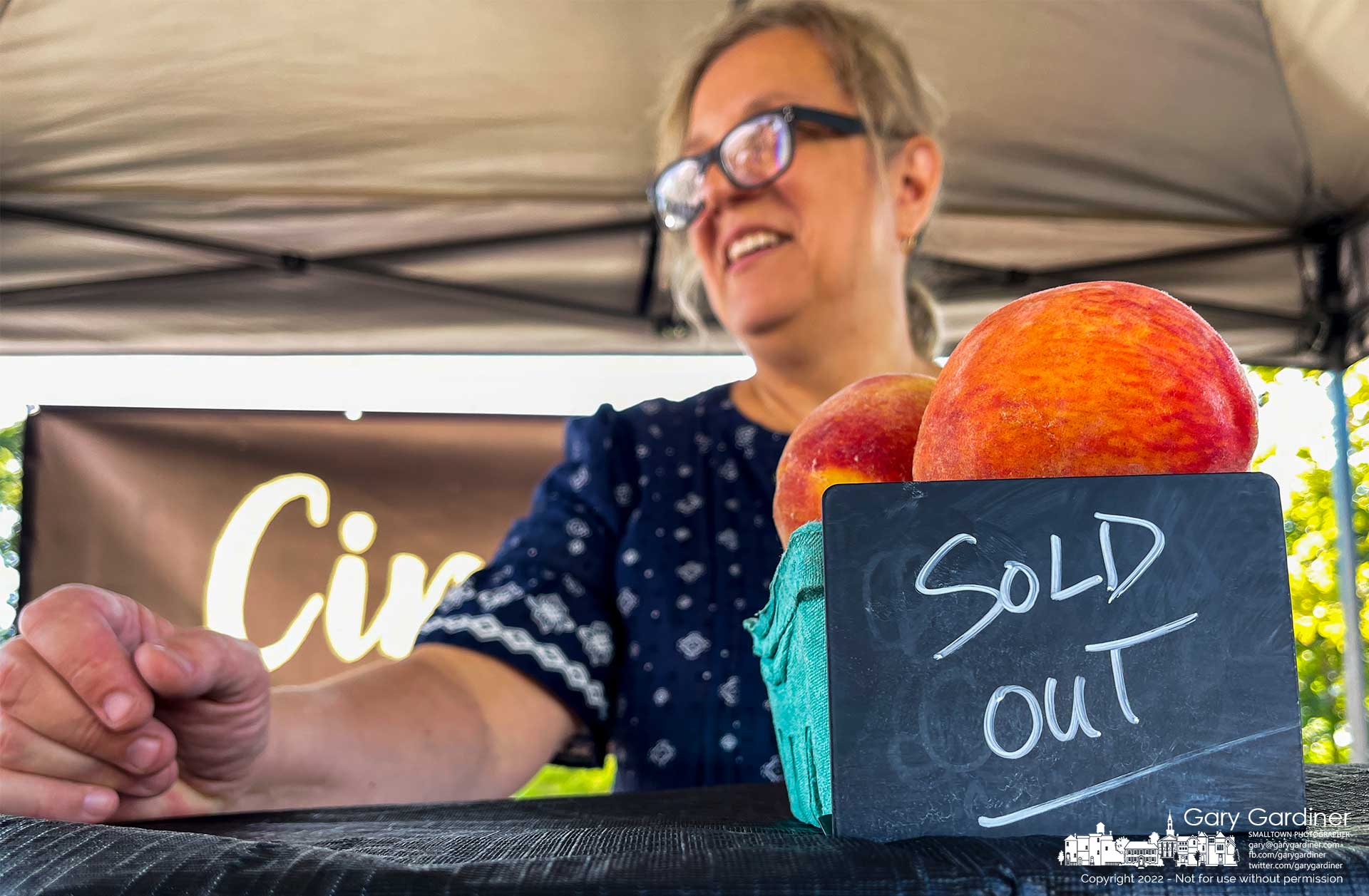 A cinnamon bun baker uses a neighborsvendors fruit and display box to hold up her "Sold Out" chalkboard less than halfway through the Saturday Farmer's Market. Several vendors sold out early with the large crowd arriving early in the morning. My Final Photo for August 27, 2022.