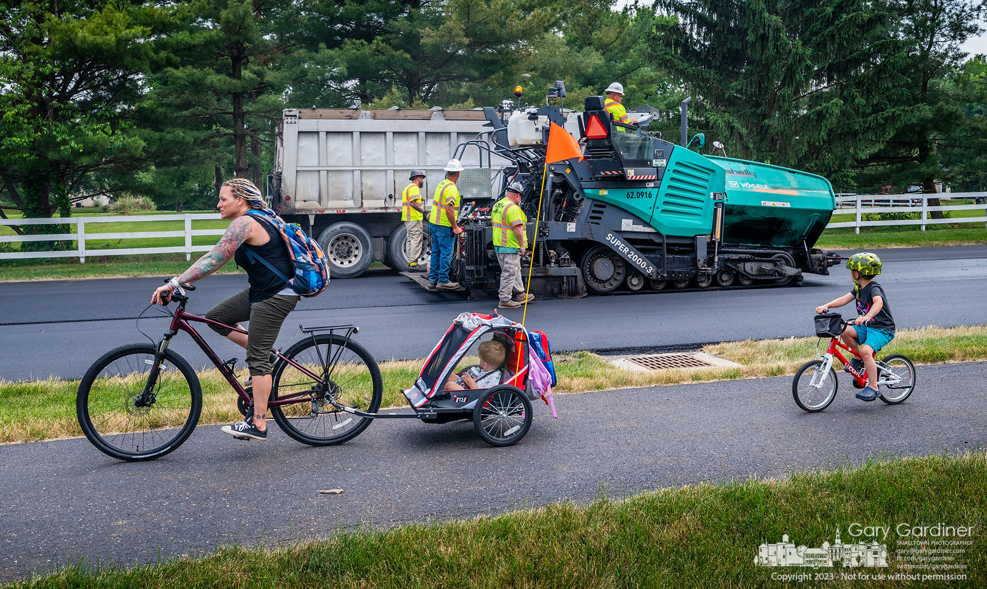 An entourage of parents and children ride their bikes past a paving company laying asphalt on Maxtown Road in Genoa Township. My Final Photo for June 8, 2023.