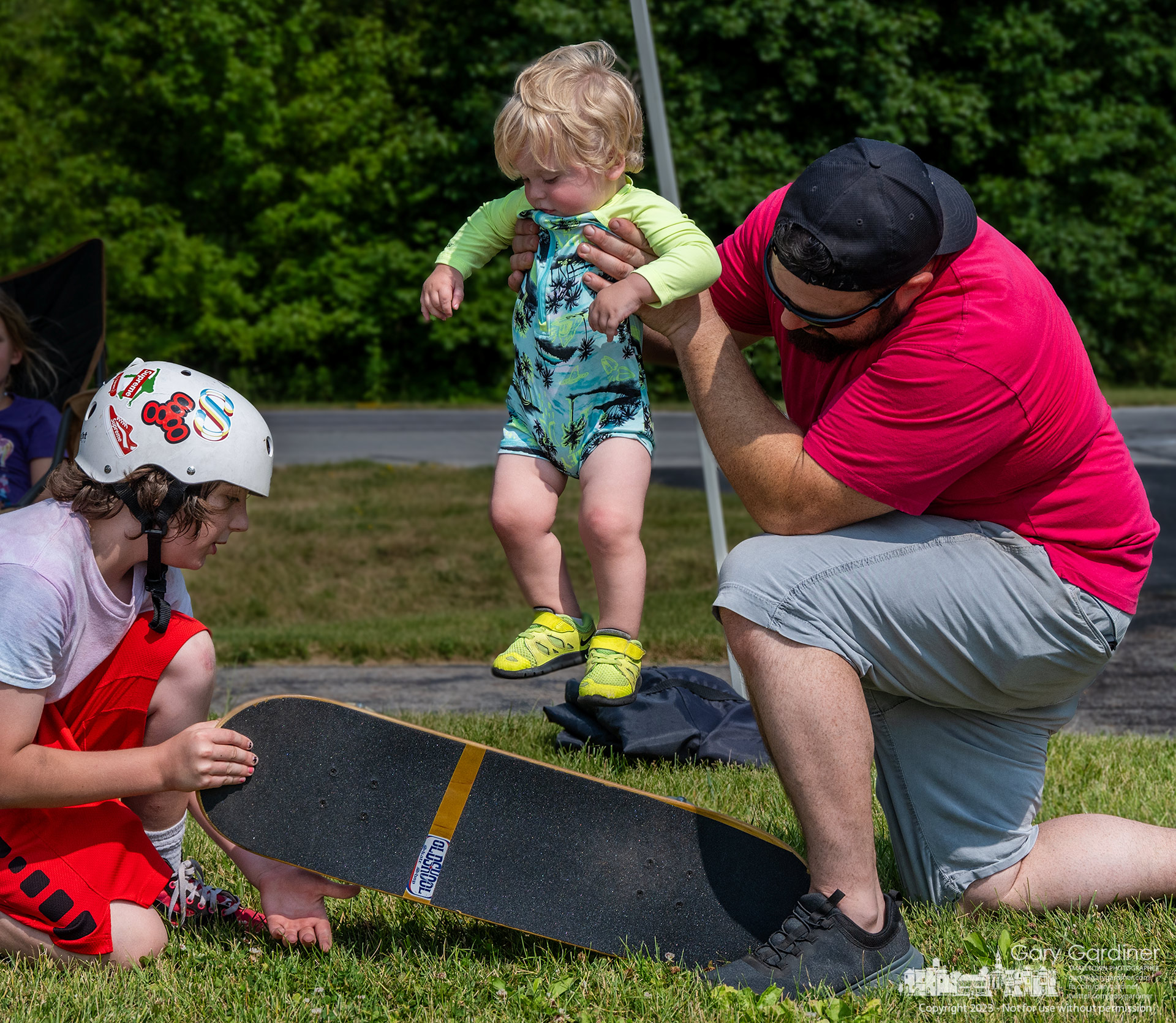 A toddler gets assistance from father and brother to learn a standard skateboarder's trick, the kick-flip, during Old Skool Skateshop's Father/Child skate around and competition Sunday. My Final Photo for June 18, 2023.