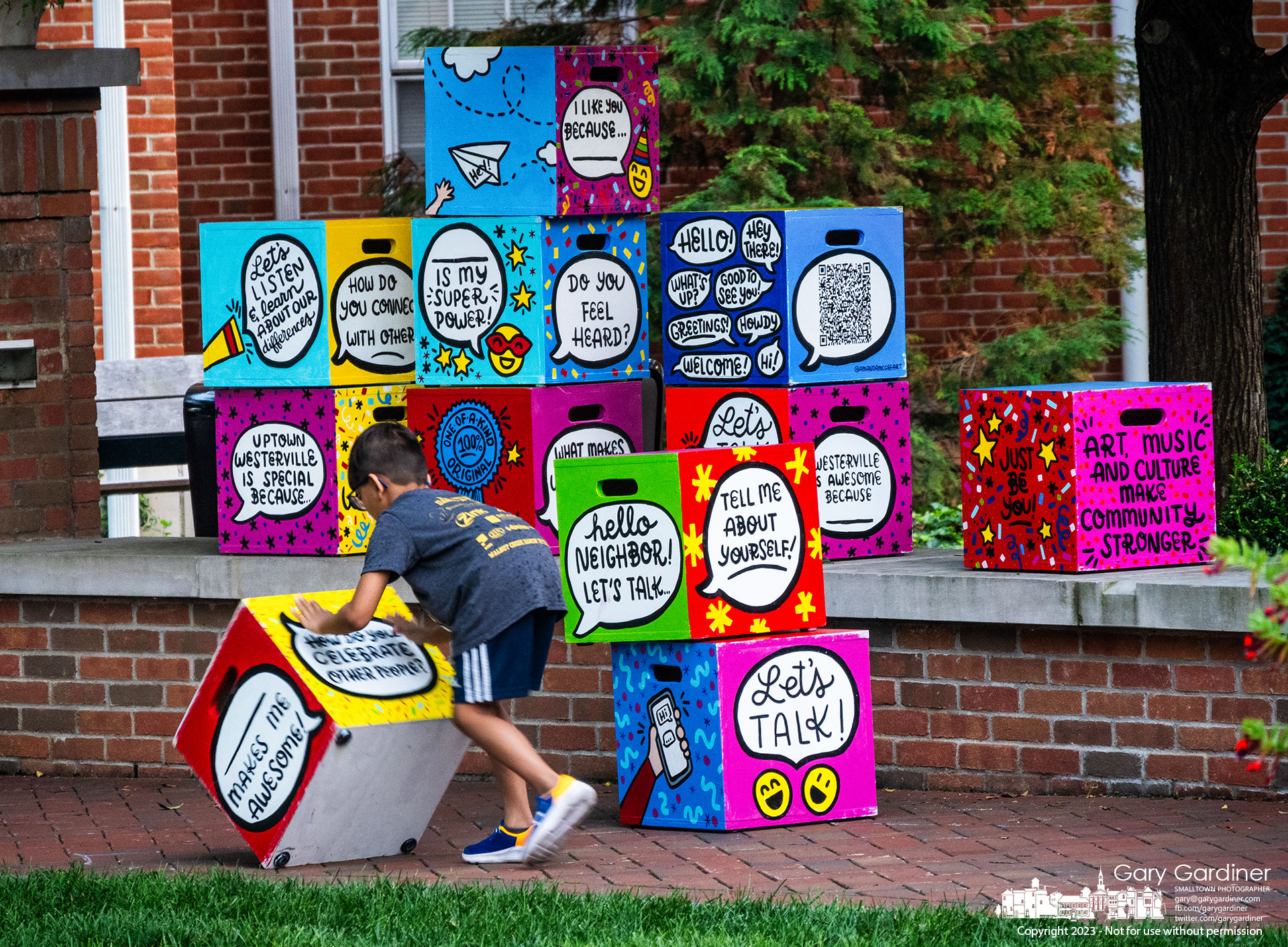 A young boy repositions one of artist Amanda McGee's Conversation Cubes designed tro be portable seats and conversation starters in the greenspace in front of City Hall. My Final Photo for August 18, 2023.