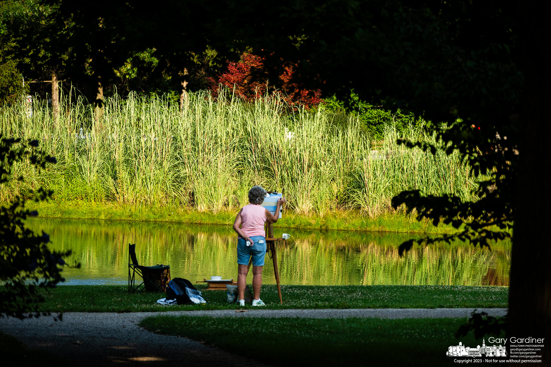 A plein air artist applies chalk pastels to her canvas as she stands in the late afternoon shadows edging over the pond at Heritage Park. My final Photo for August 3, 2023.