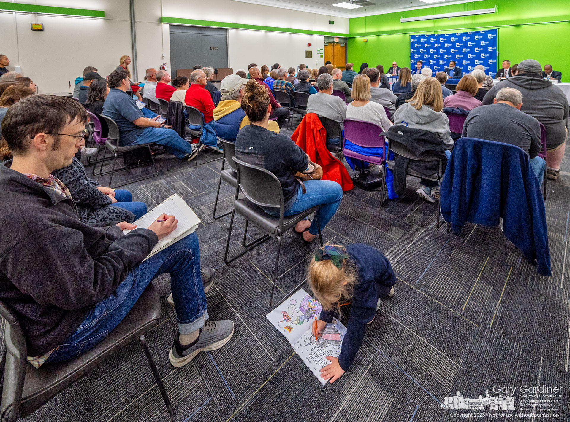 A toddler keeps herself busy coloring between the lines of her coloring book as a man in the audience takes notes during the Chamber of Commerece City Council Candidate Forum at the library. My Final Photo for October 9, 2023.