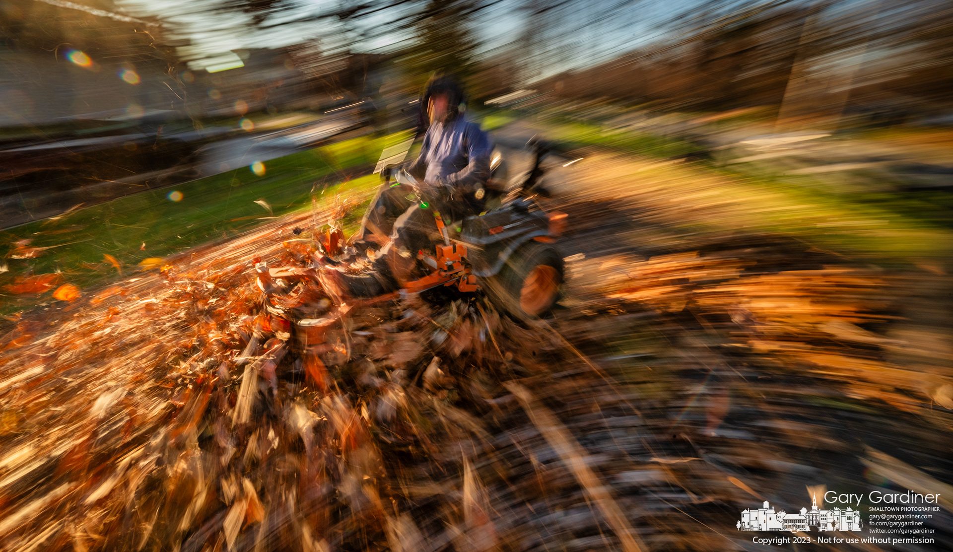 A landscaper uses a large lawnmower with its deck lowered to move thick piles of leaves off grass and sidewalk along Maxtown Road to a collection point for removal. My Final Photo for November 29, 2023.