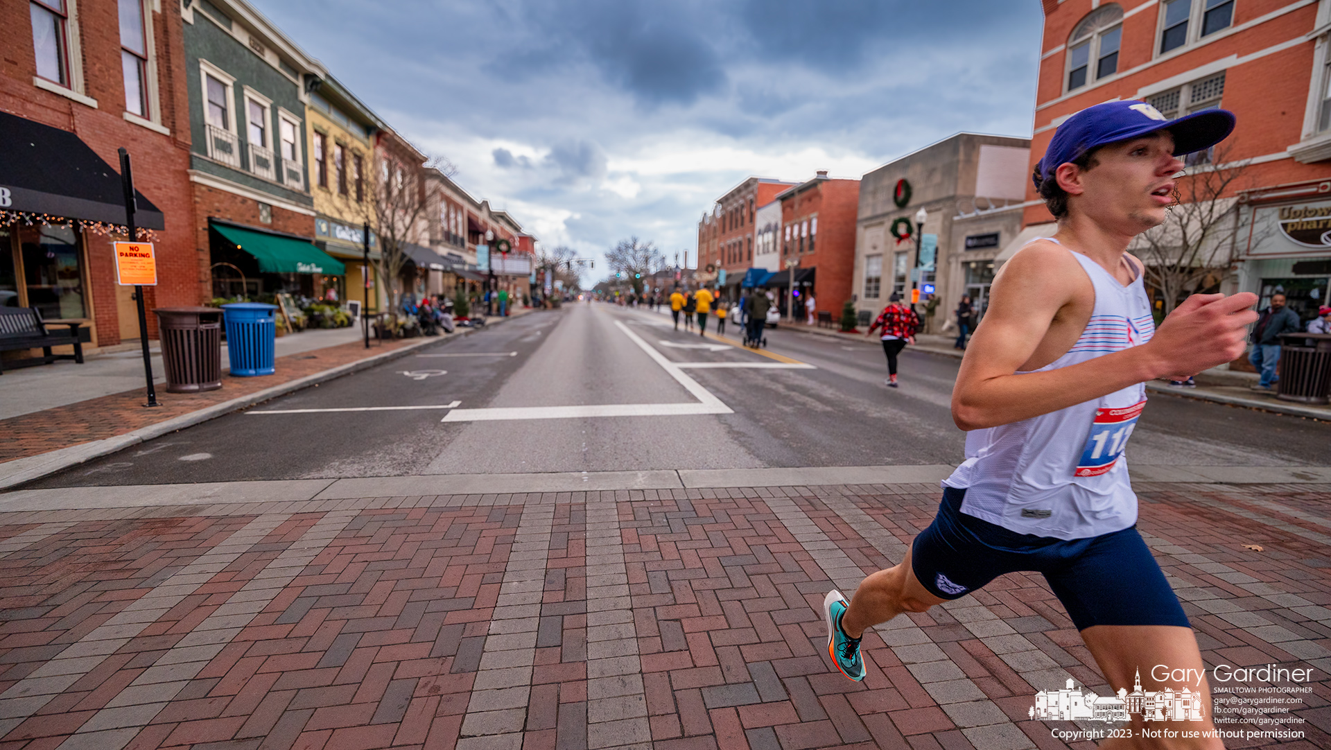 The first racer to cross the finish line at the Rudolph Run 5K crosses State and Main in Uptown Westerville on the return loop of the course as other runners reach about one-fifth of the racecourse on the first loop. My Final Photo for December 3, 2023.