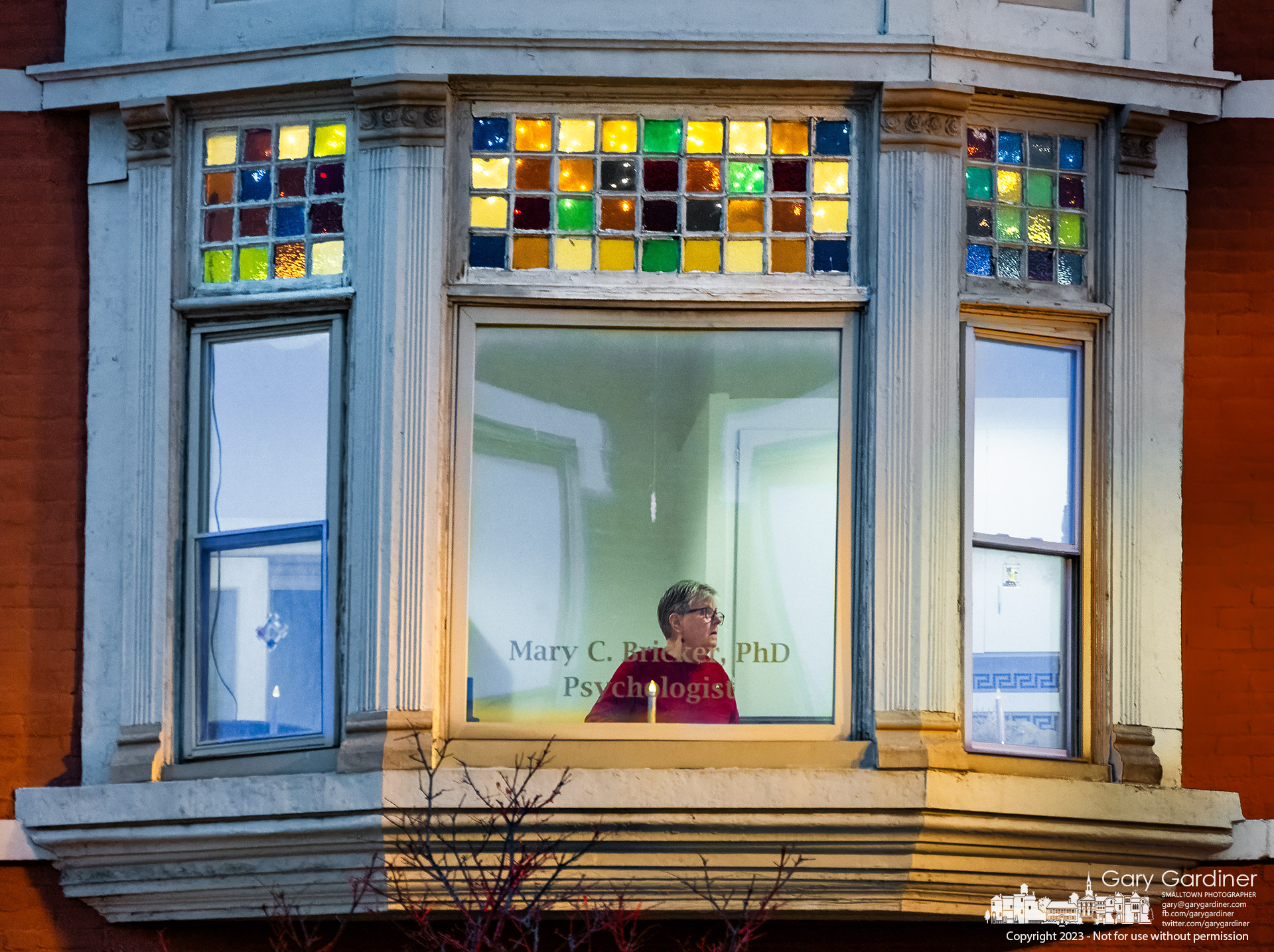 Psychologist Mary Bricker stands at one of the windows in her second-floor offices in the Holmes Hotel where the stained-glass portions of her windows are illuminated with lights during the Christmas season. My Final Photo for December 13, 2023.