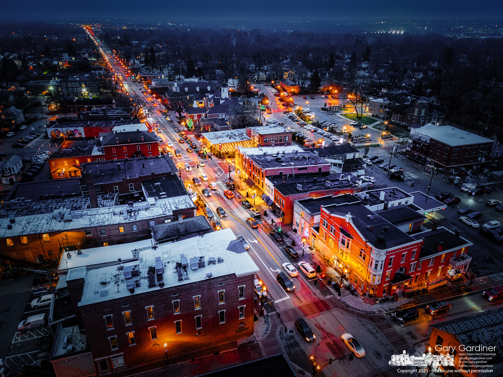 Late afternoon traffic travels along State Street in Uptown Westerville where the city glows in the darkness of heavy cloud cover less than half an hour after sunset. My Final Photo for December 27, 2023.