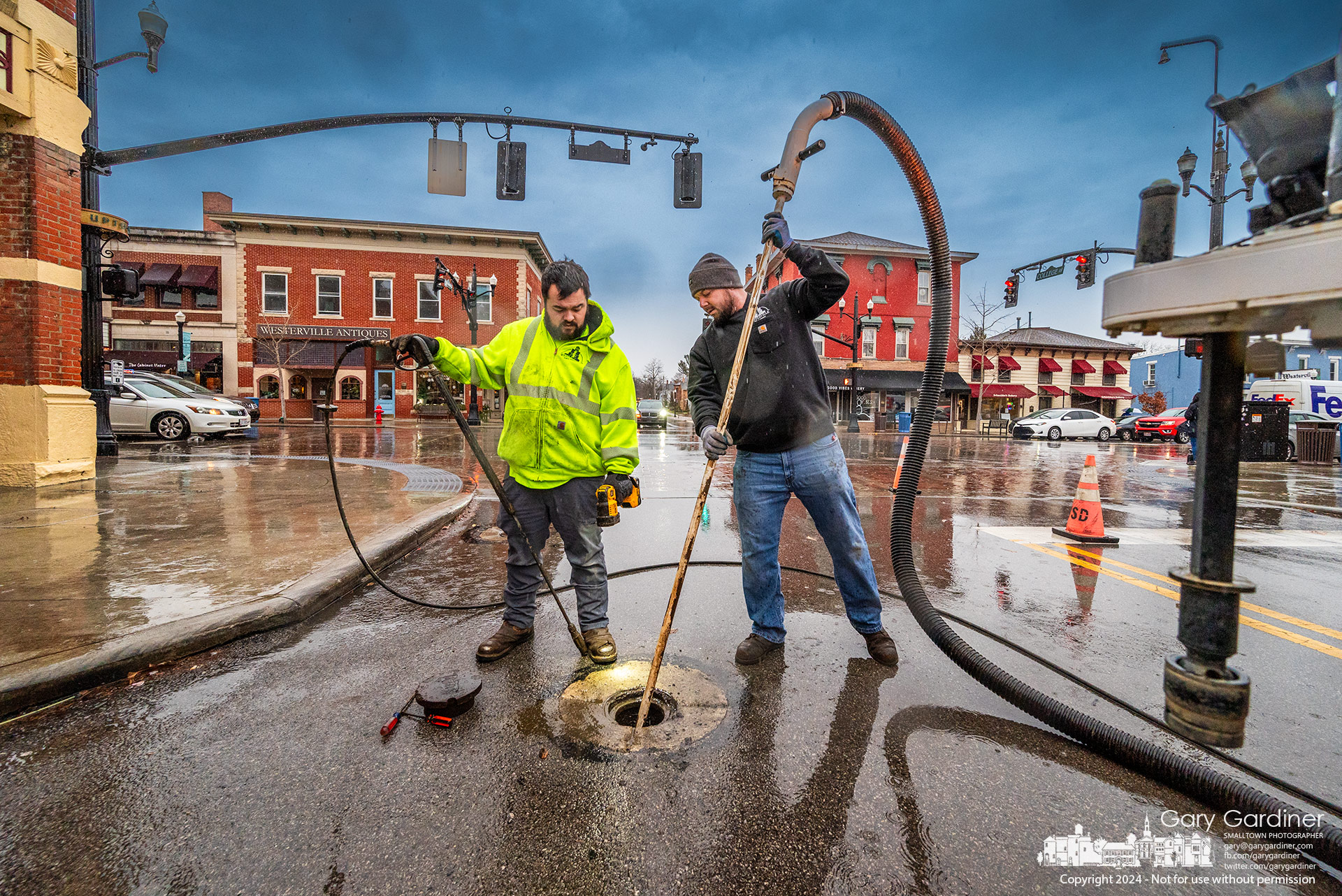 City workers stand in the rain as they power wash and vacuum clear a water valve on West College at State Street as part of the regular cleanup of service valves in city streets. My Final Photo for January 12, 2024.