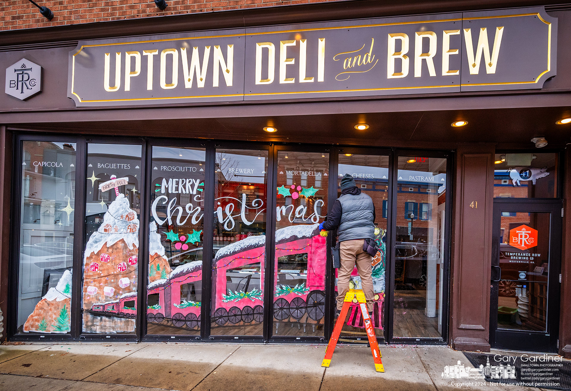 Micah Crockett removes the decorative Christmas painting on the seven door panels at the front of Uptown Deli and Brew on Epiphany, marking the Christmas season's end. My Final Photo for January 6, 2024.