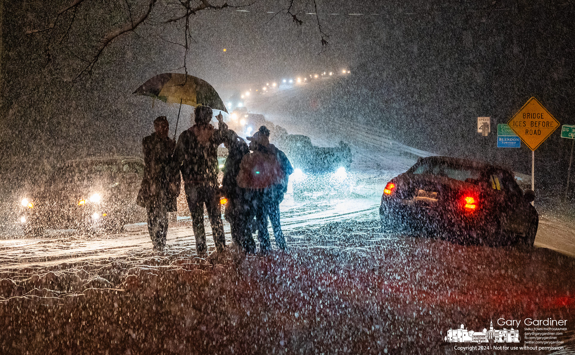 A line of westbound cars sits idle on Central College at the bridge below Hoover Dam waiting for police to open the roadway following a series of car crashes without injury during the evening snowfall. A group of drivers, passengers, and parents of drivers stand together waiting for tow trucks to remove their vehicles. My Final Photo for February 16, 2024.