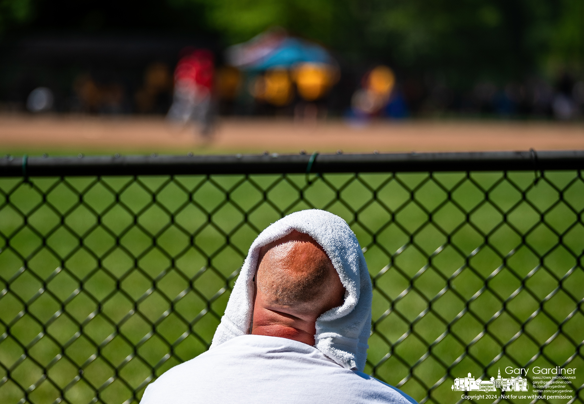 A youth baseball fan partially shields himself from the full sun from his seat behind the left field fence during a rain-delayed makeup game for WYBSL at Huber Ridge Park. My Final Photo for May 19, 2024.