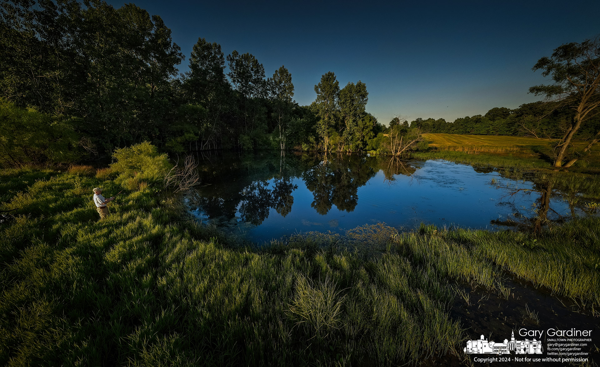 Mark Glenn casts for large-mouth bass in a pond secluded behind farm field and shrubs, with the water reflecting the fading light, hoping for a good catch as the sun sets behind him. My Final Photo for June 24, 2024.