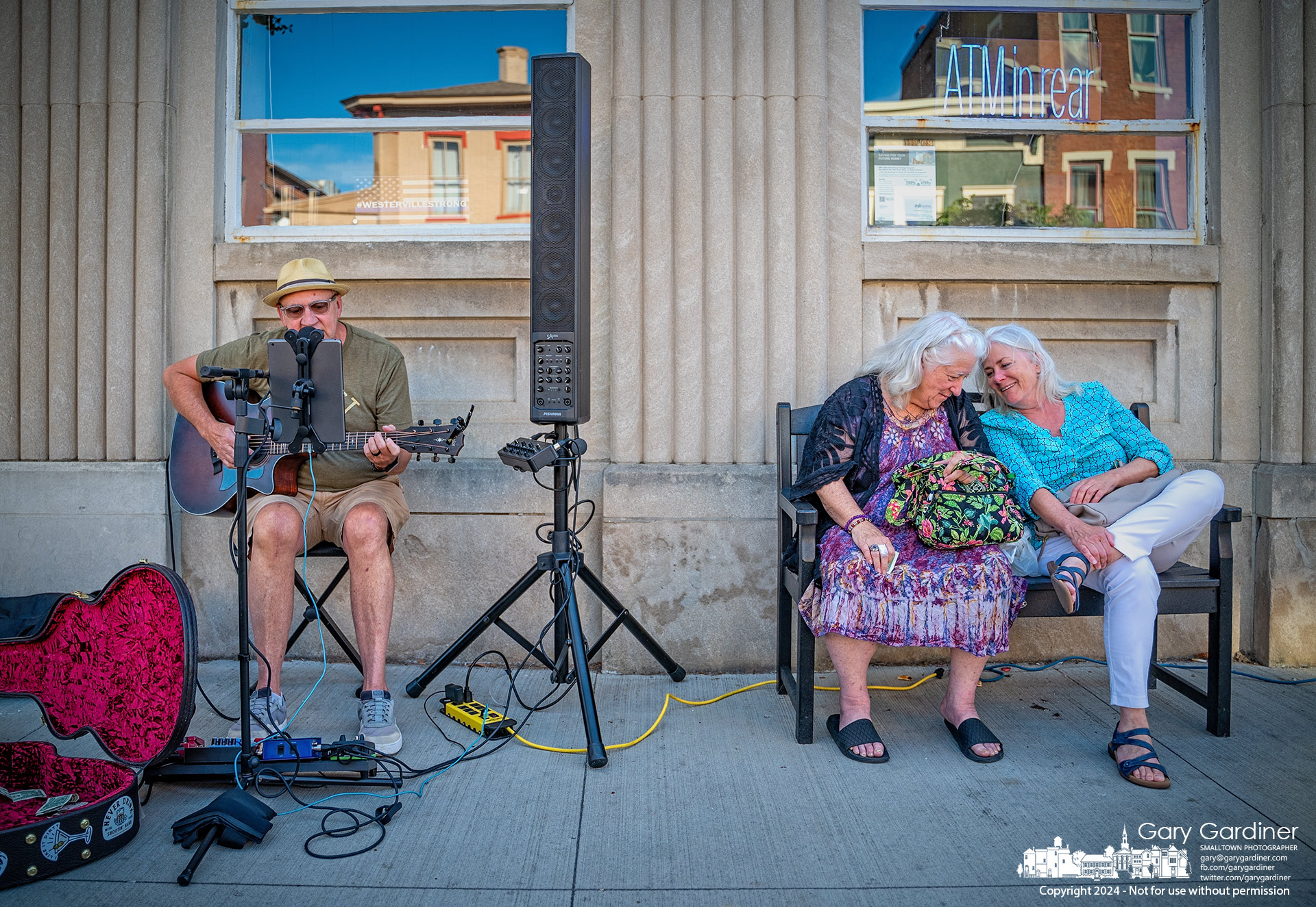 Jim Meadows performs in front of Middlefield Bank in Uptown Westerville Friday night where two women mistakenly identified as his groupies enjoy a laugh between themselves. My Final Photo for July 19, 2024.