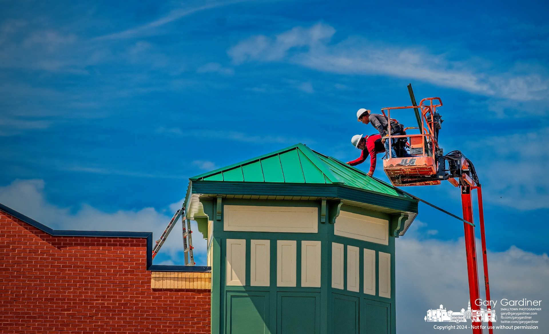 Roofers lean out of their bucket lift to install ribbing joining section of the metal roof atop the turret at the multi-use building under construction on West College in Uptown. My Final Photo for June 2, 2024.