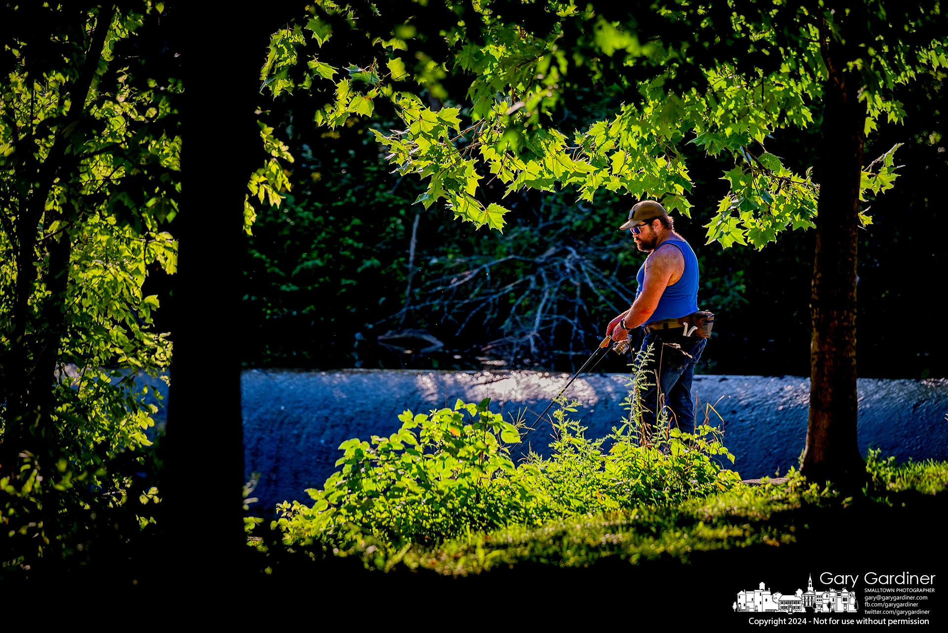 A fisherman standing in the late afternoon light at Alum Creek North Dam is framed by trees and bushes along the creek's edge. My Final Photo for August 11, 2024.