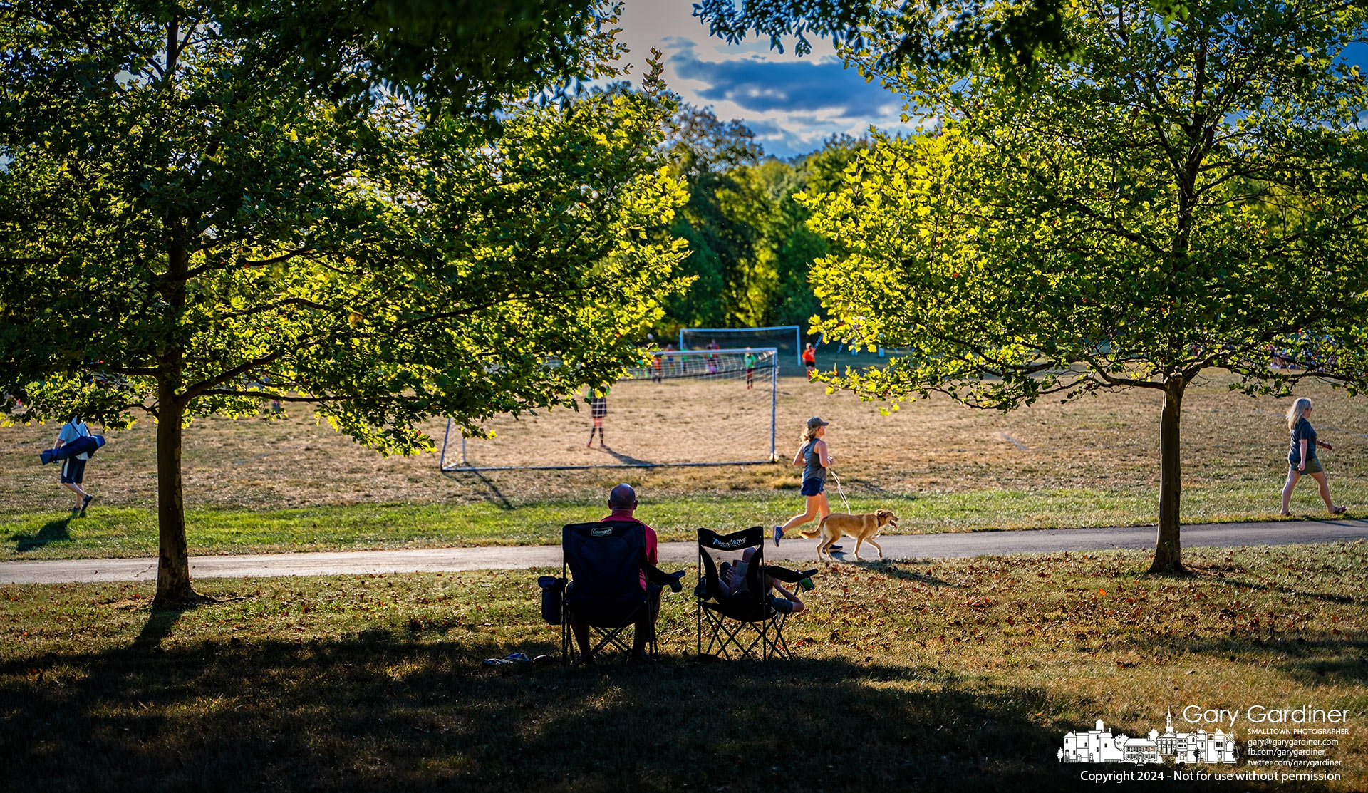 A young boy and man sit in the shifting afternoon shade, watching a soccer match on the Sports Field at the Community Center as others run, walk, and play along the path and fields. My Final Photo for September 18, 2024.