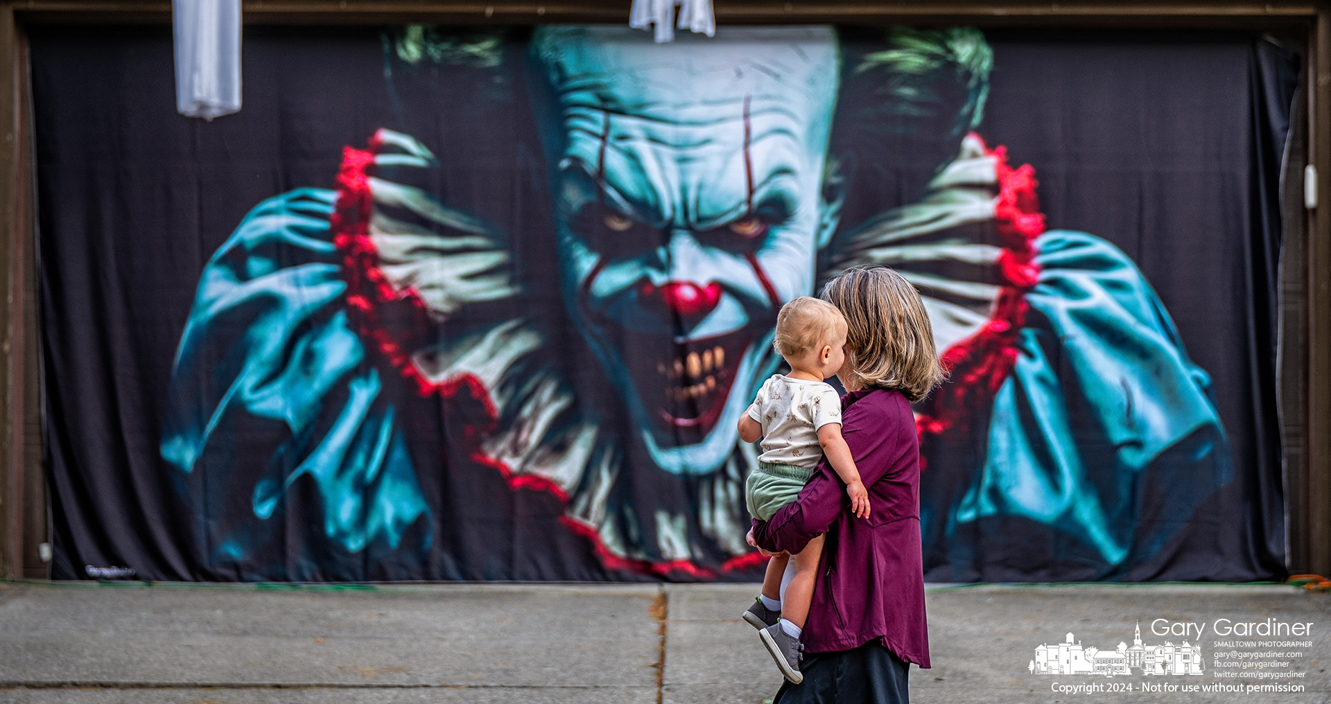 Mather and child casually walk by a garage door covered with a Pennywise the Dancing Clown mural as part of a macabre Halloween decoration on Spring Road. My Final Photo for October 23, 2024.