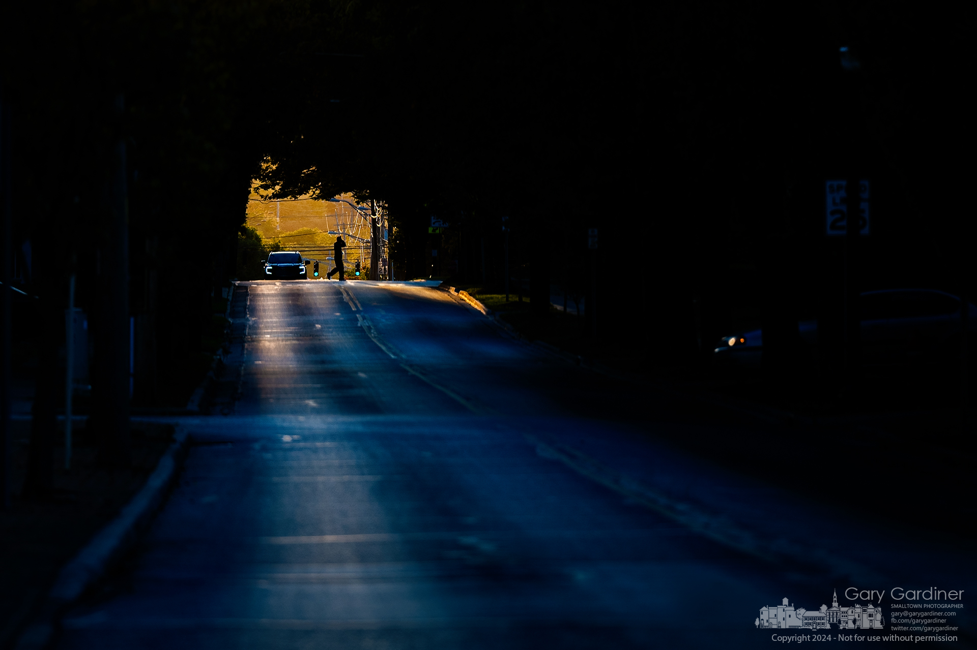 A lone pedestrian crosses West Main Street through deepening shadows on the quiet street, illuminated by the golden light of the setting sun spilling over the horizon. My Final Photo for October 11, 2024.