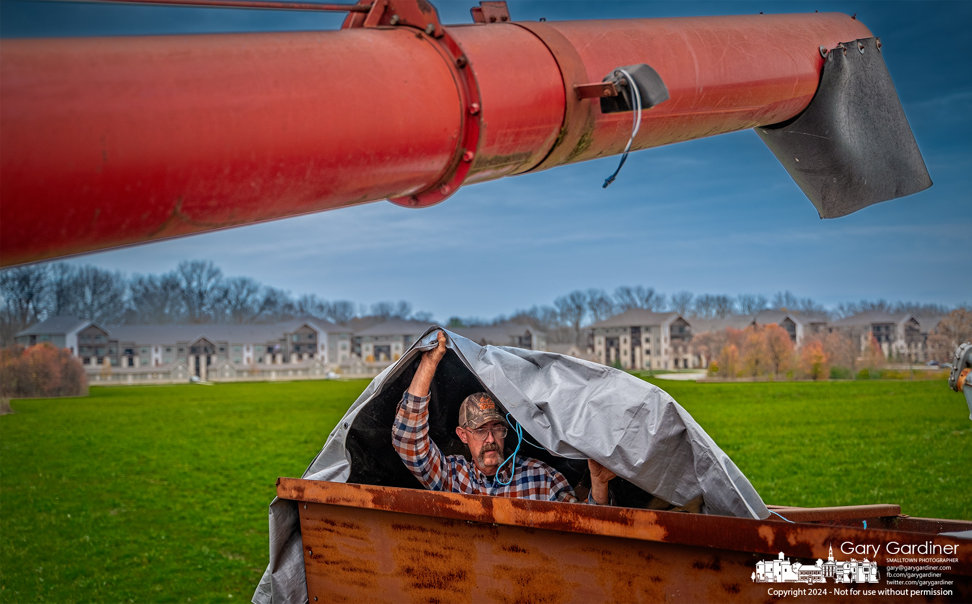 xFarmer Rodney Parker pulls a tarp over his head to cover a partially filled grain wagon after a brief afternoon shower dampened the soybeans on the Braun Farm just enough to make them unharvestable, forcing an end to harvest until the rain ends and the fields again are dry. My Final Photo for November 18, 2024.