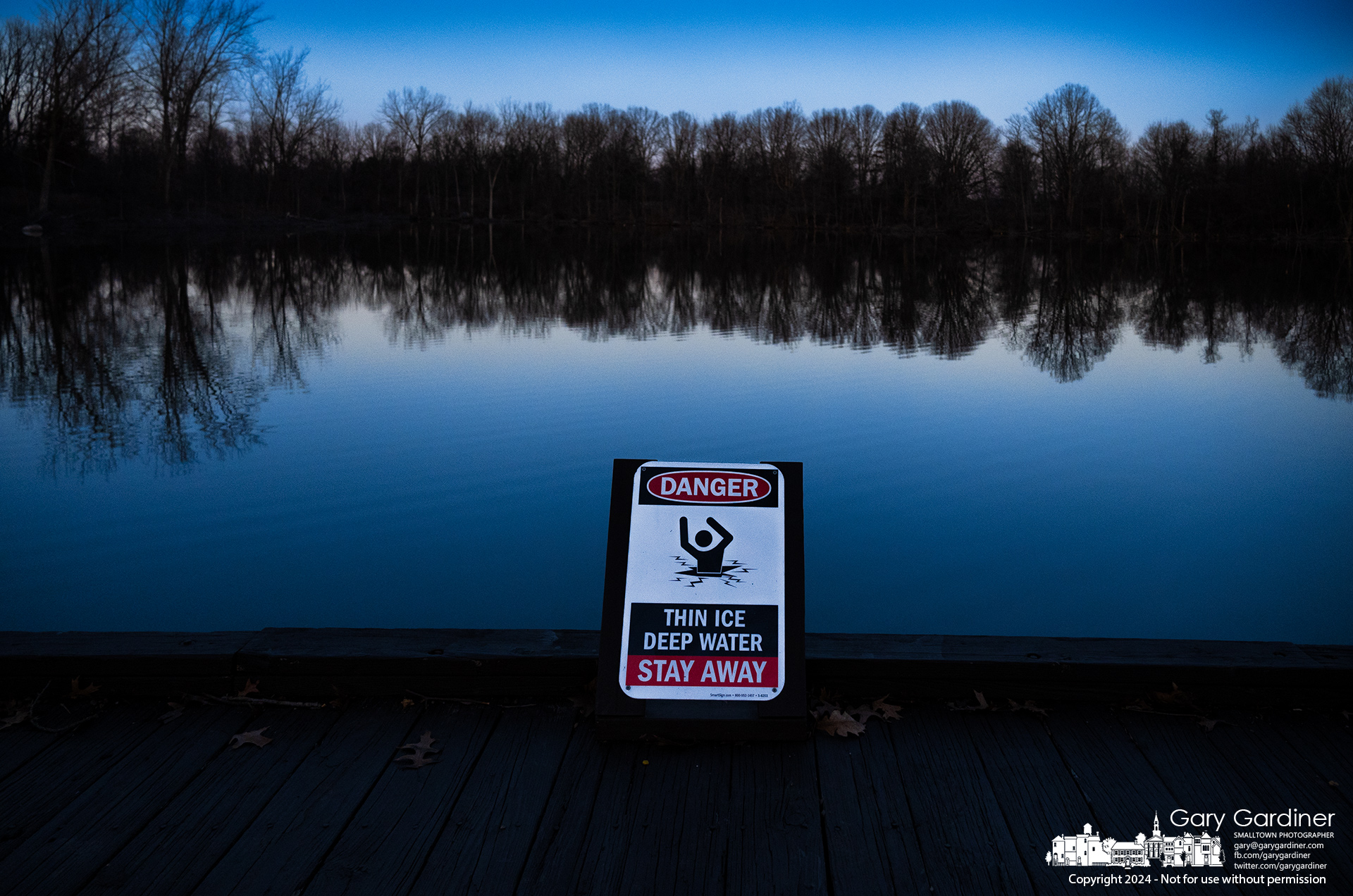 The smooth waters of Schrock Lake sit in a blue glow after sunset with the thin ice warning sign that might be more of a weather forecast and a statement of conditions as a series of polar storms are forecast to bring Arctic temperatures and winds to the Midwest. My Final Photo for December 30, 2024.