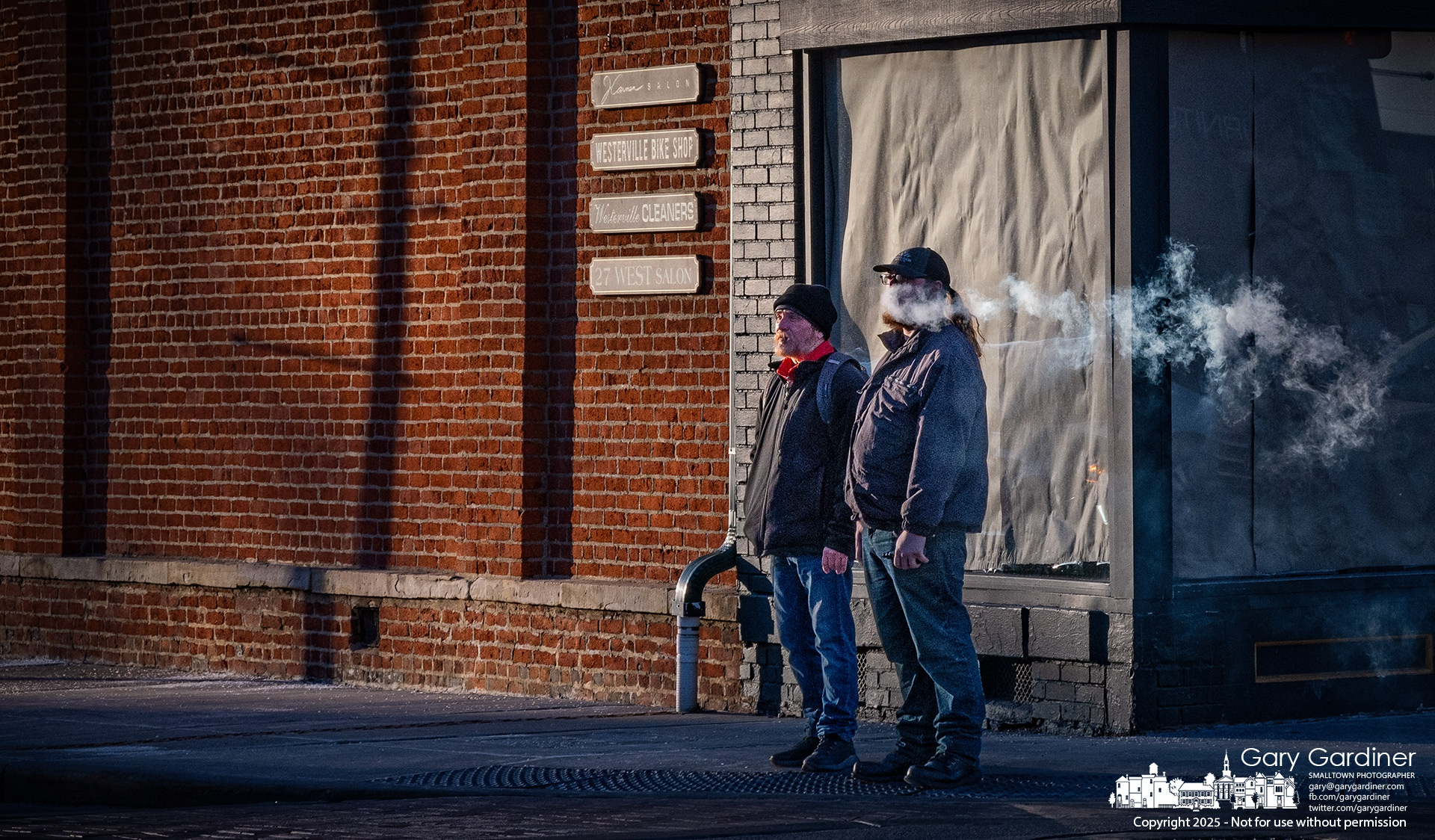 Two men waiting to cross Main Street stand in light from the setting sun and a strong southwesterly wind. My Final Photo for January 28, 2025.