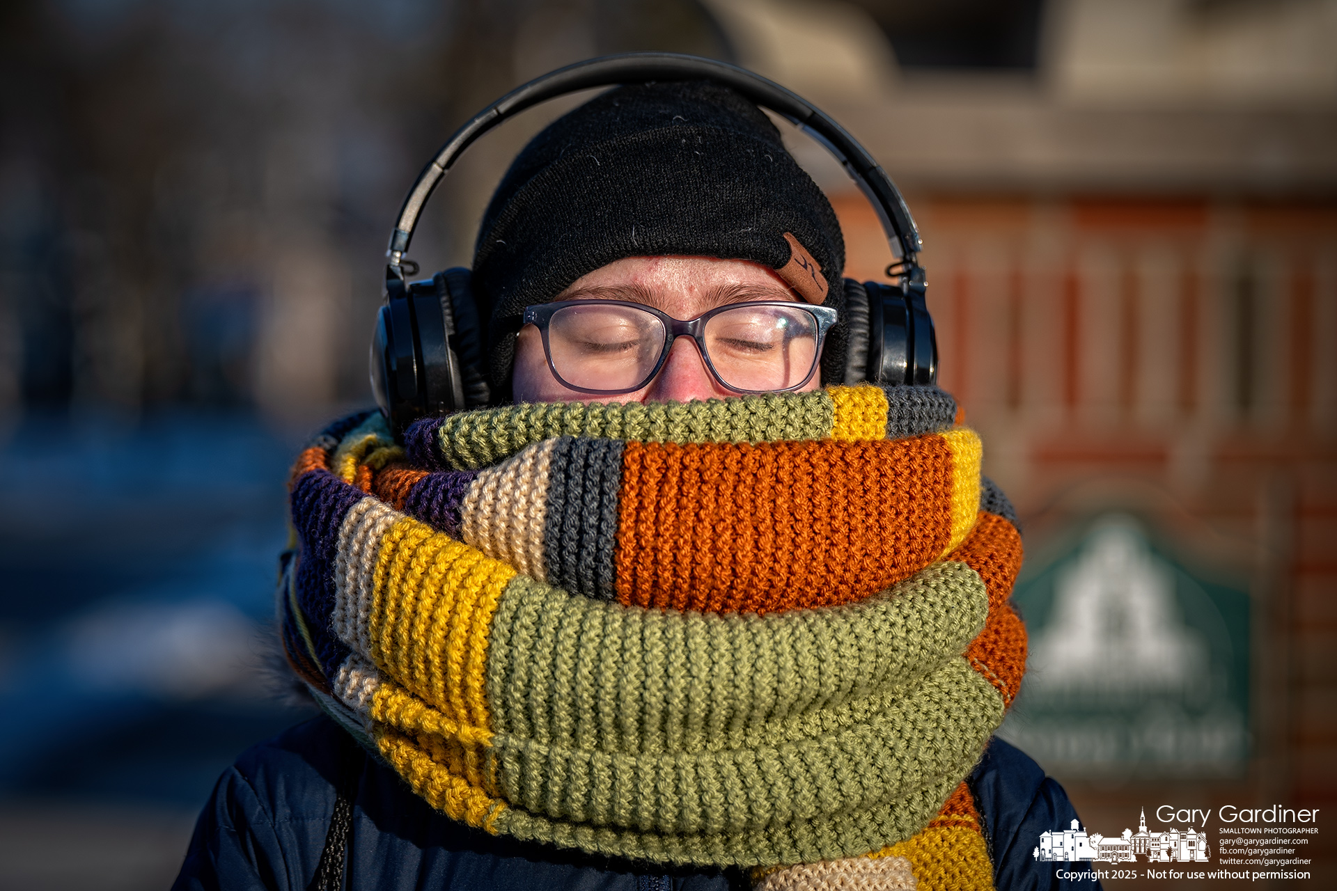 Theodore Marshall closes their eyes as they wait to cross State Street in the slight warmth of the setting sun, using earphones for music and ear protection, as well as a heavy scarf to protect against single-digit temperatures in Uptown Westerville. My Final Photo for January 15, 2024.