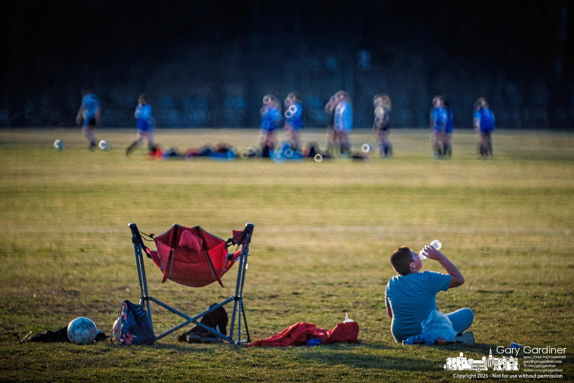 A soccer player takes a water break before returning to a pickup game with friends while some of his family members practice with their team in the background. My Final Photo for March 10, 2925.