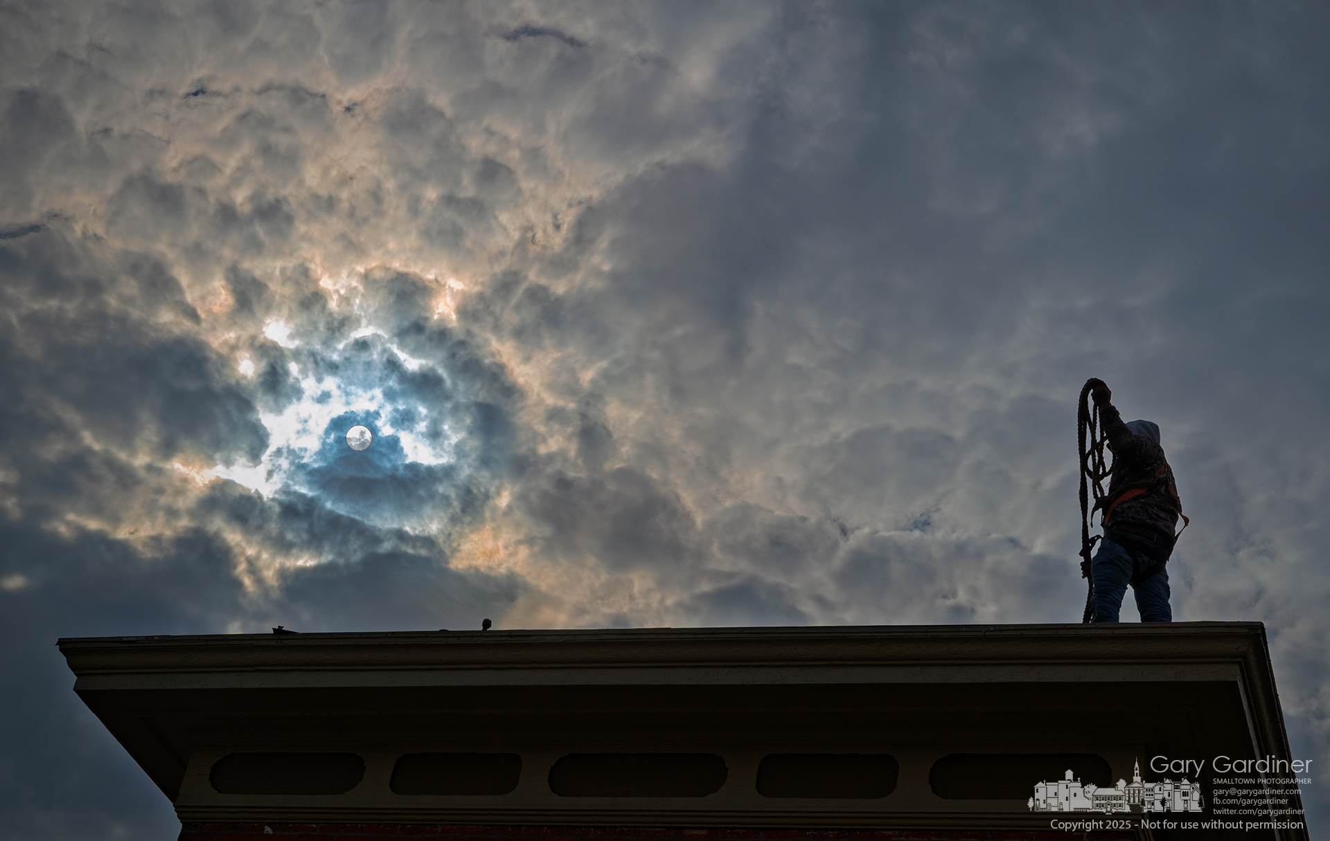 A roofer checks his safety gear before shifting positions to lay shingles under a cloudy sky. As he moves, the clouds part, revealing the afternoon sun. My Final Photo for March 3, 2025.