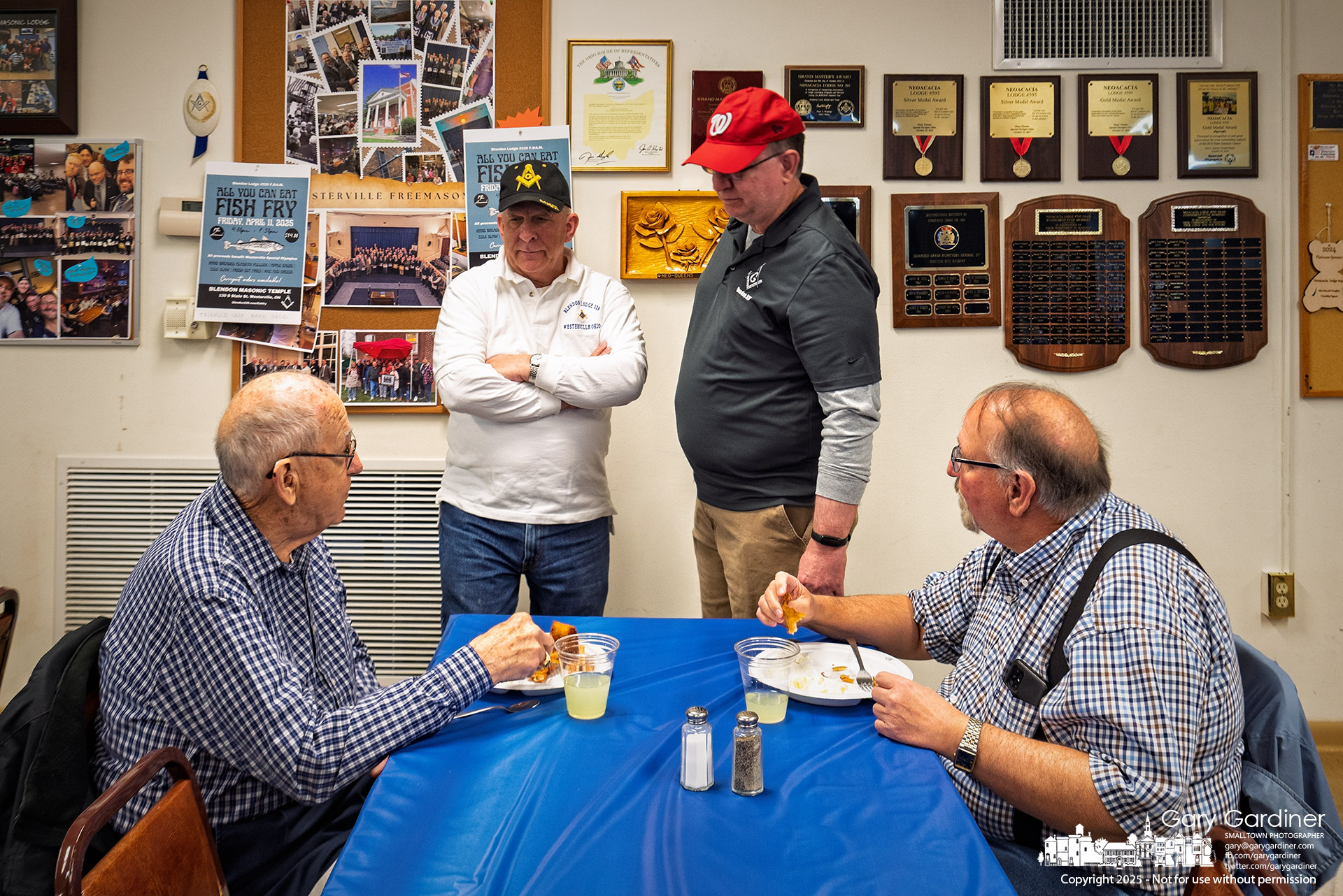 At the Masonic Hall’s annual fish fry, two men sit eating as two others arrive to join them, framed by a wall of plaques and awards that echo the years of service, friendship, and tradition behind the gathering. My Final Photo for April 11, 2025.