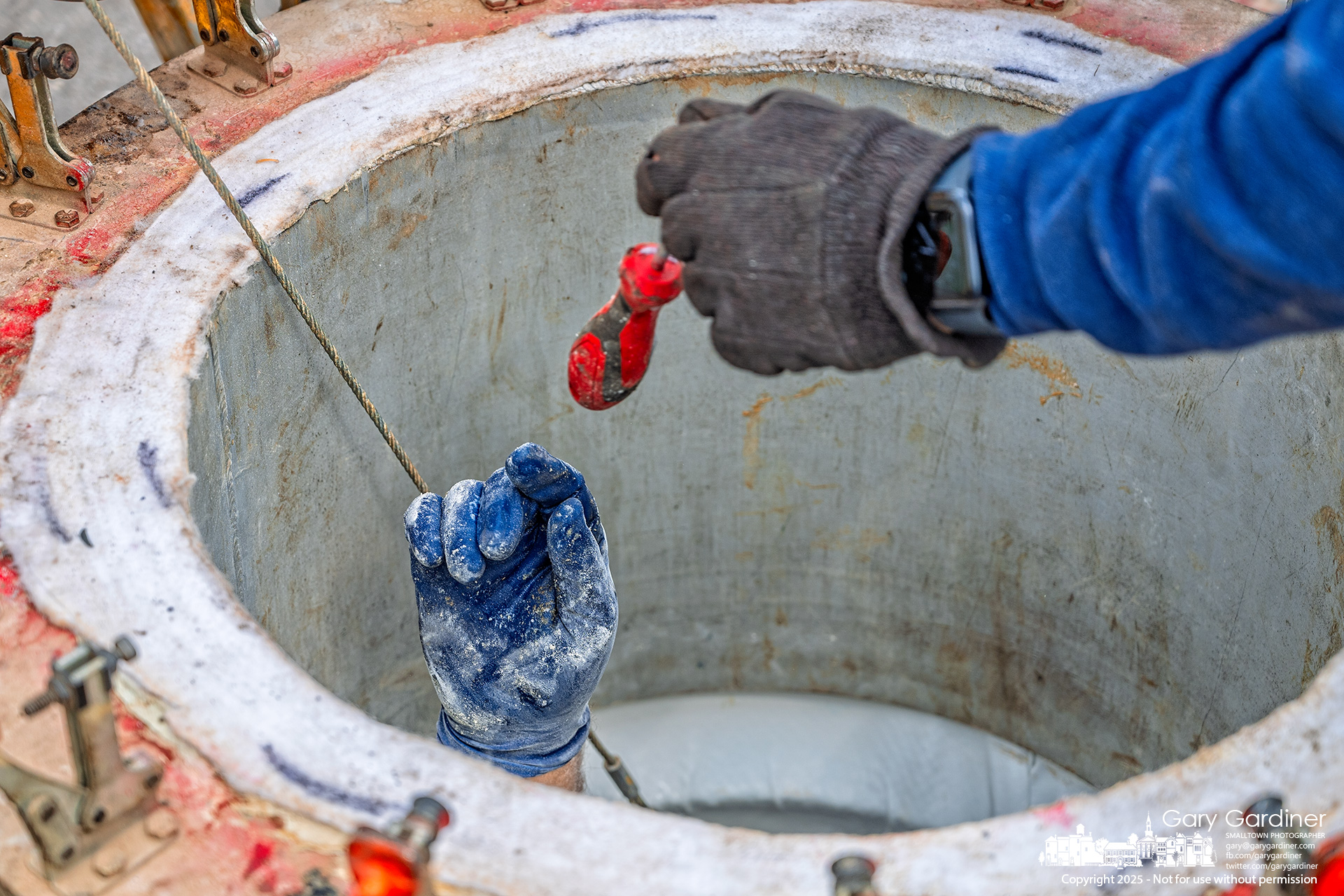 A gloved hand reaches from inside a sanitary sewer for a screwdriver to complete the installation of a liner in the manhole. My Final Photo for April 9, 2025.