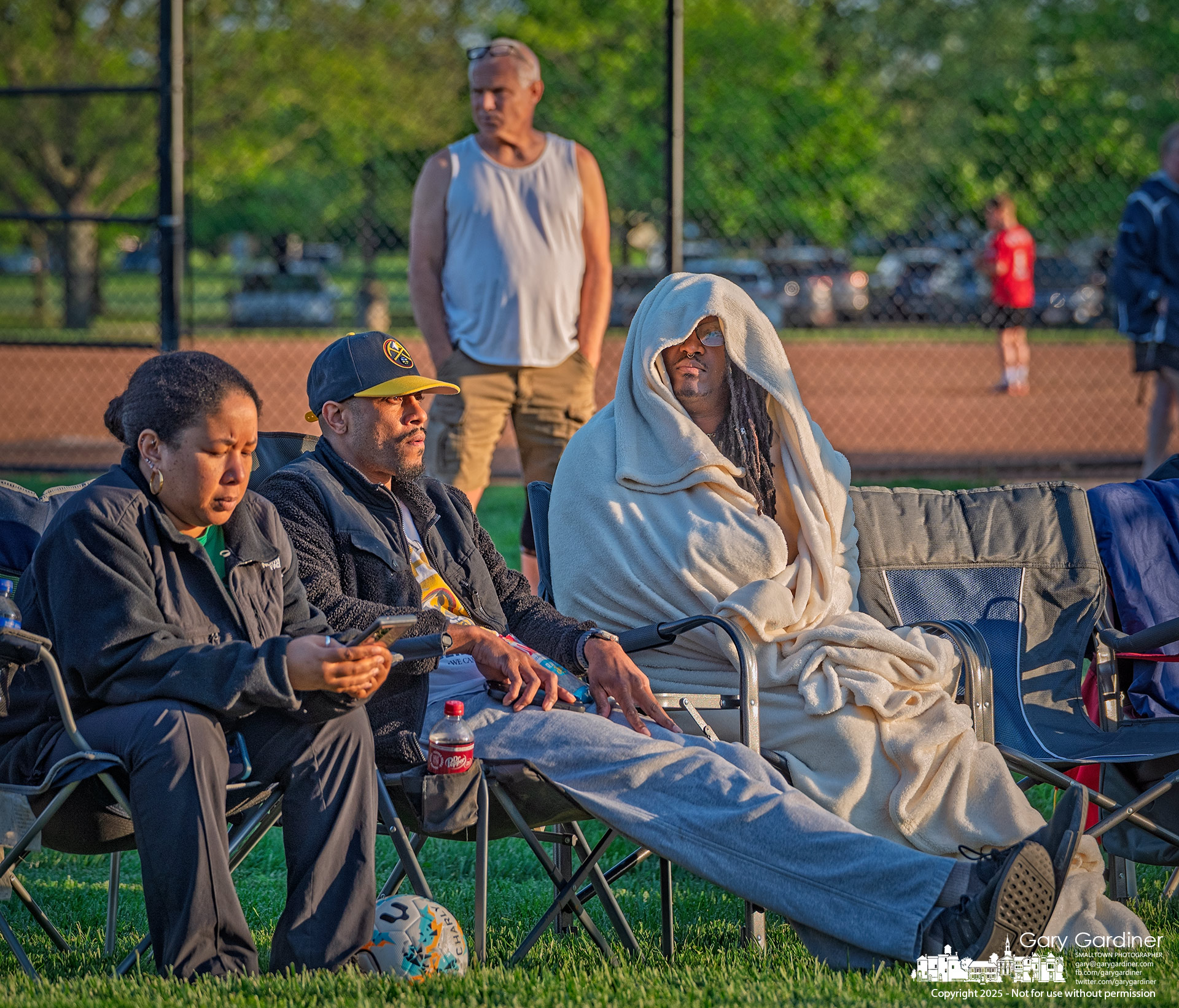 A spectator bundled in a large blanket shields himself from the evening chill, while another fan stands comfortably in a sleeveless shirt during a youth baseball game at Huber Village Park. My Final Photo for May 8, 2025.