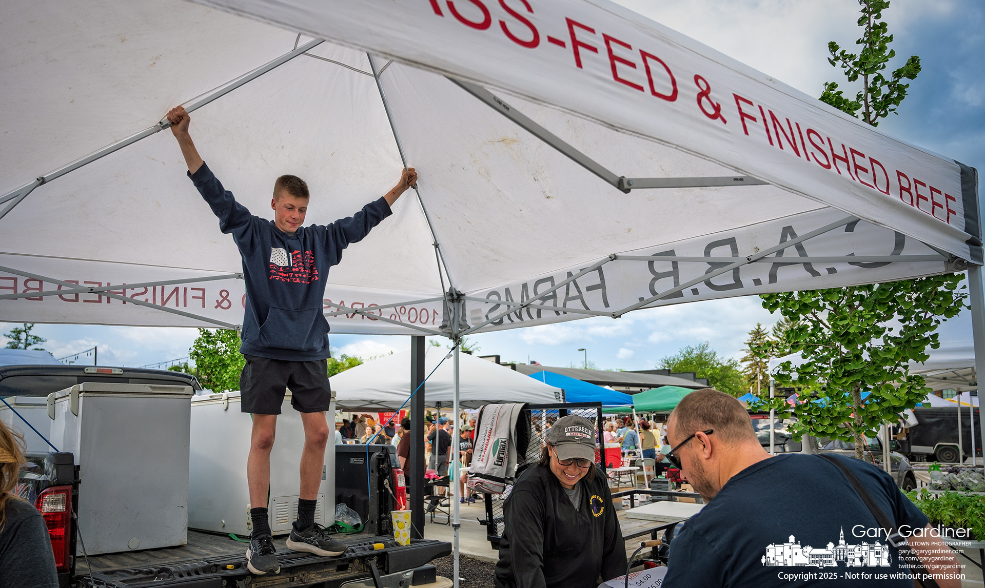 A C.A.B.B. farmhand lends a hand by holding down the farm’s tent during gusty winds at the Saturday Farmers Market—the first day of the season at its new location in the COhatch and North High Brewing parking lot on South State Street. My Final Photo for May 17, 2025.