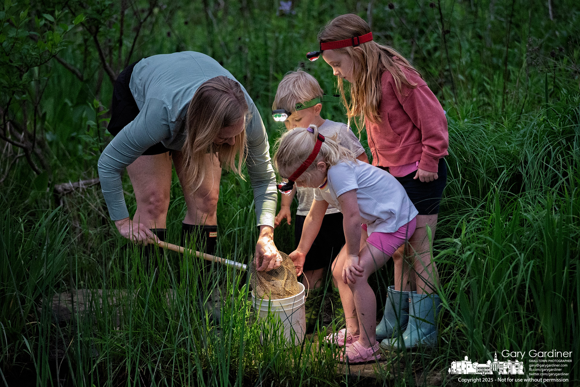 A mother looks on as children use their headlamps to examine tadpoles found during Frog Friday at the Highlands wetlands. My Final Photo for May 16, 2025.