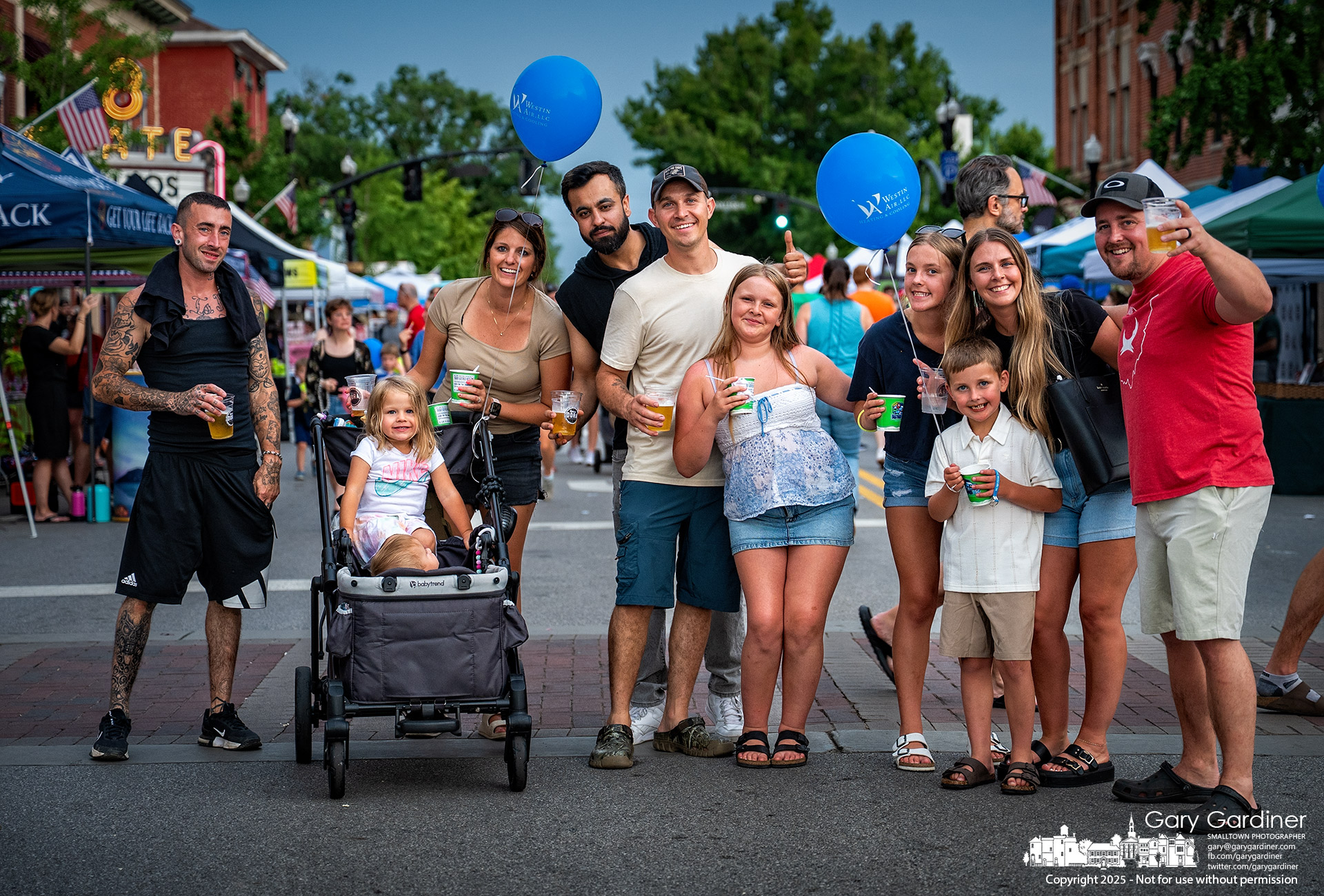 Friends and family gather at the crossroads of State and Main Streets in Uptown Westerville during the Fourth Friday festival, pausing to request their portrait for Take My Picture!, a 50-year photographic series capturing community life at the invitation of those photographed. My Final Photo for June 27, 2025.