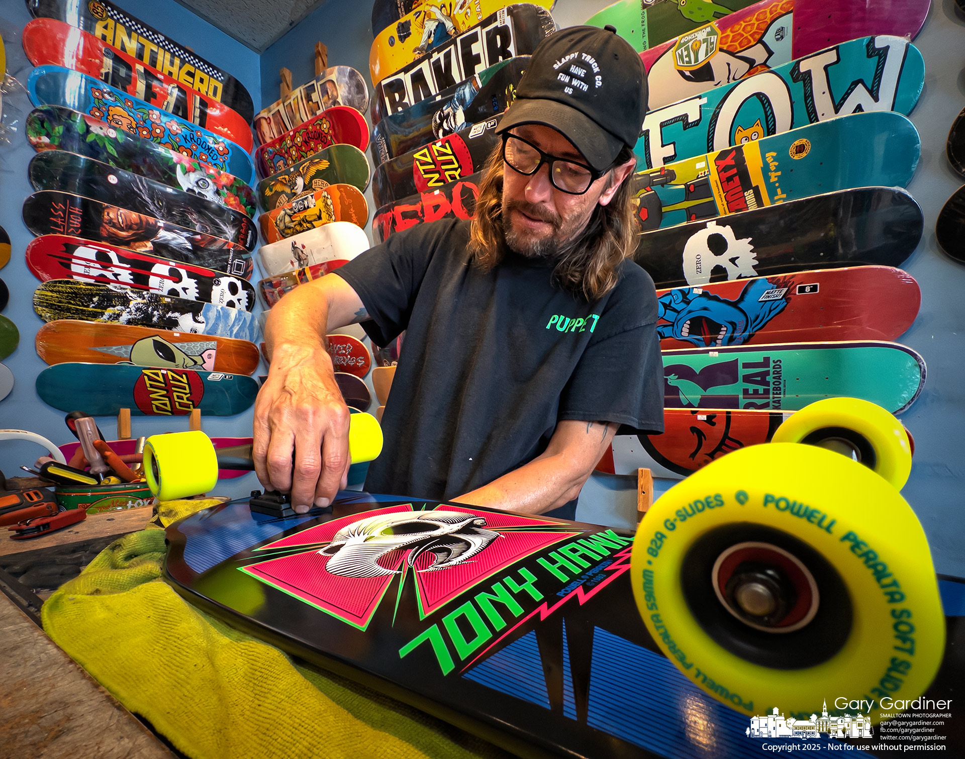 Wally Carl tightens the trucks on a Tony Hawk cruiser at OldSkool Skate Shop, where walls lined with bold, colorful decks set the tone for a hands-on approach to skateboarding culture for 24 years. My Final Photo for June 21, 2025.
