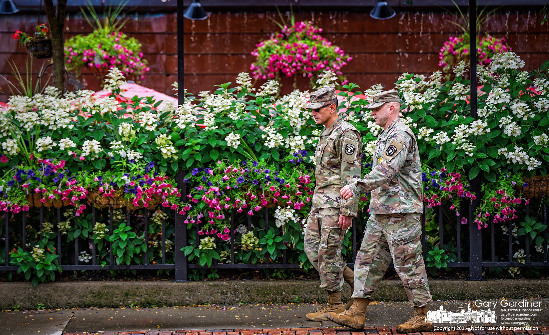 Camouflaged Army recruiters stand out against the vibrant flowers on the patio at Jimmy V’s on West College in Uptown. My Final Photo for July 10, 2025.