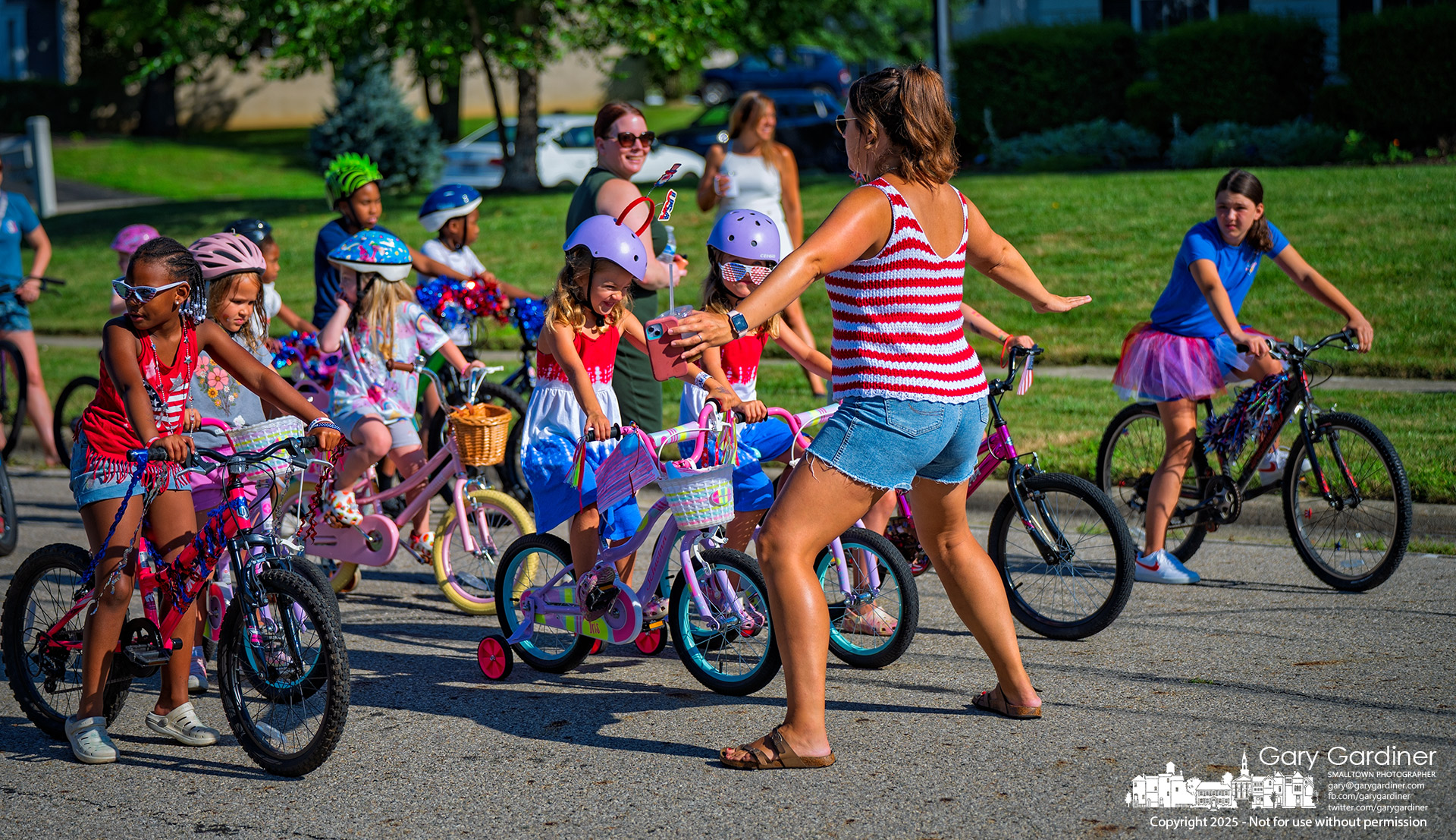 A mother steps in front of a parade of young bicyclists, signaling where they should stop at the start of the neighborhood’s July 4th bike parade on Springview Drive. My Final Photo for July 4, 2025.