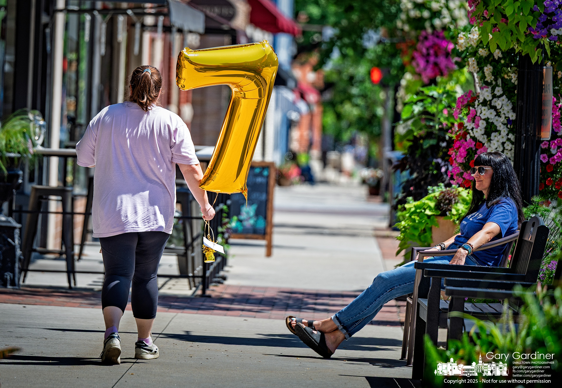 Carrying a gold number seven balloon, a woman walks through late-afternoon sunlight in Uptown Westerville as another relaxes on a bench surrounded by summer blooms. My Final Photo for July 22, 2025.