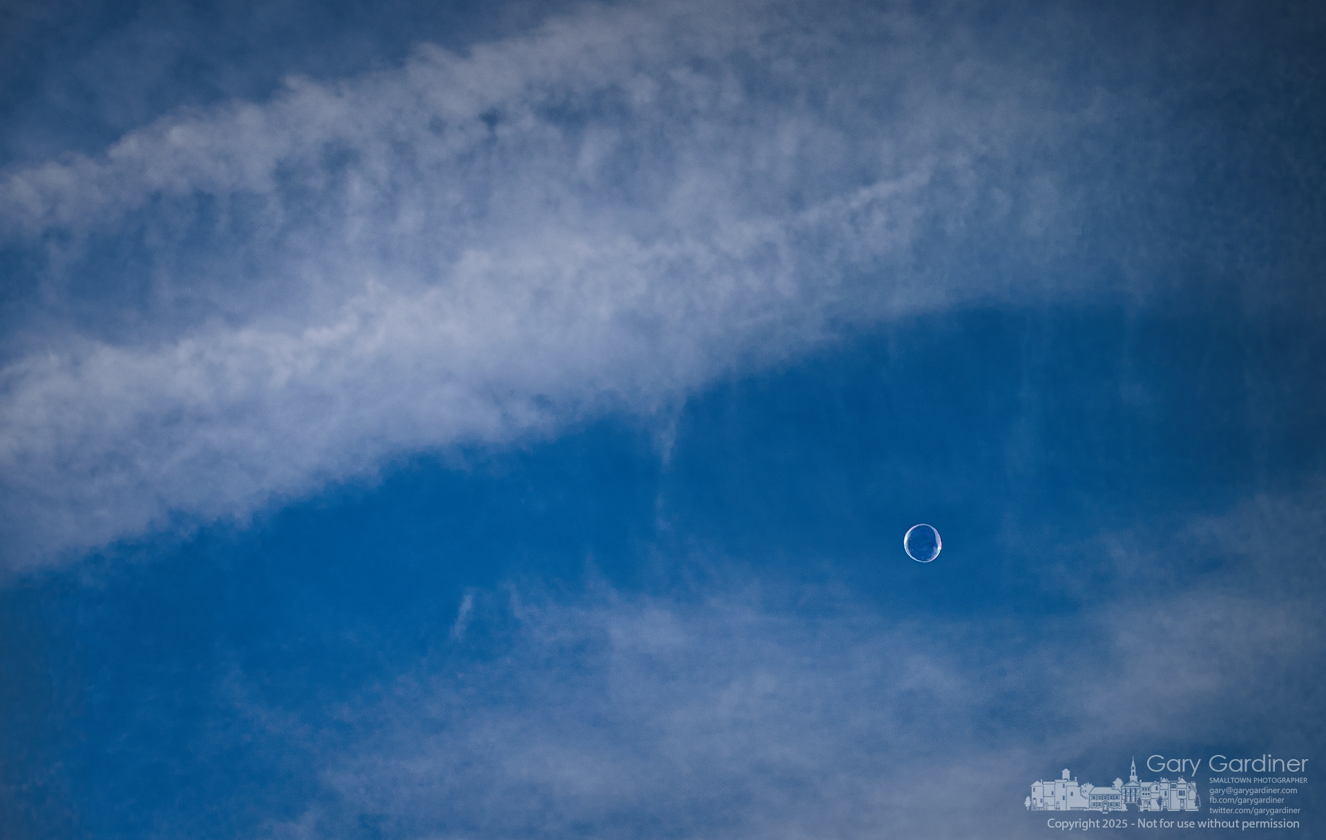 A lone two-foot soap bubble drifts across the sky above the Saturday Farmers Market, the last remnant from The Great Bubble Show as it floated beyond the crowd. My Final Photo for August 2, 2025.