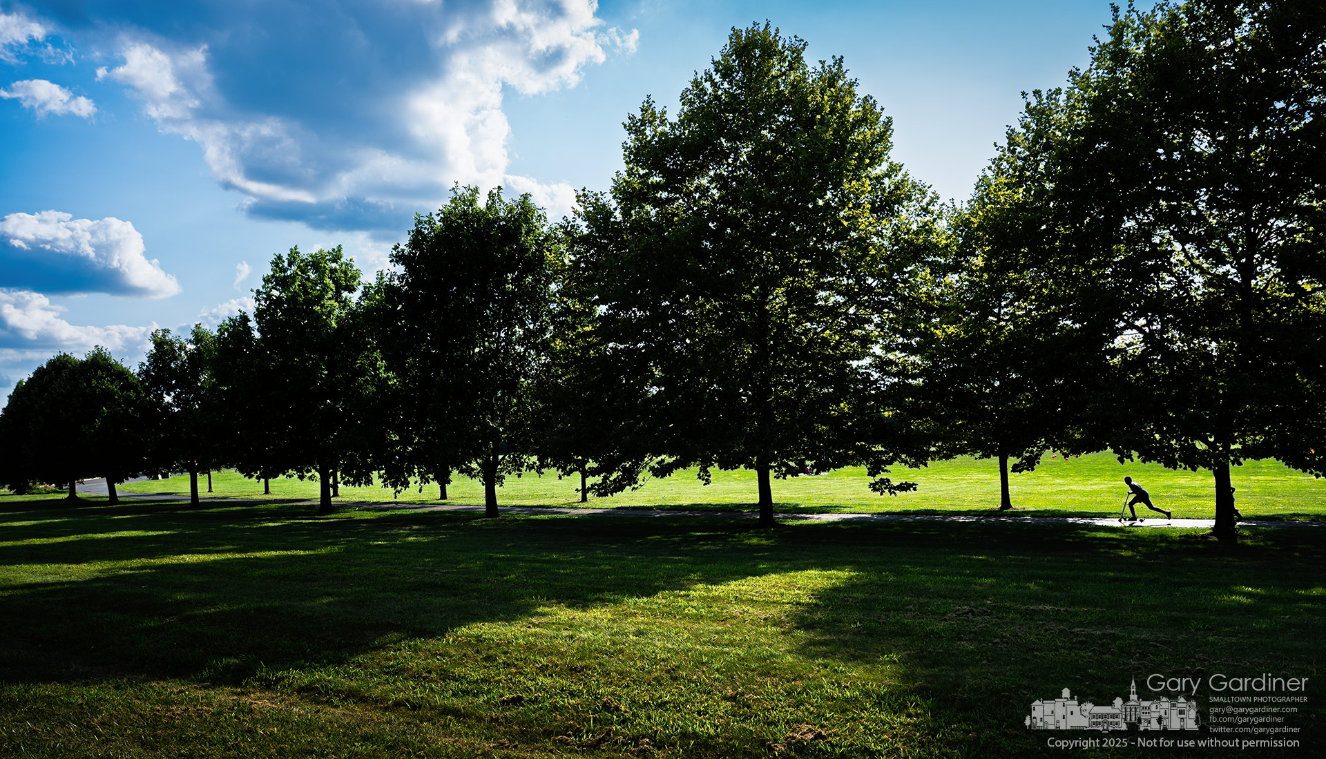 A lone scooter rider glides along a shaded path at the Westerville Sports Complex, framed by deep shadows and sunlit fields beneath towering clouds. My Final Photo for August 3, 2025.