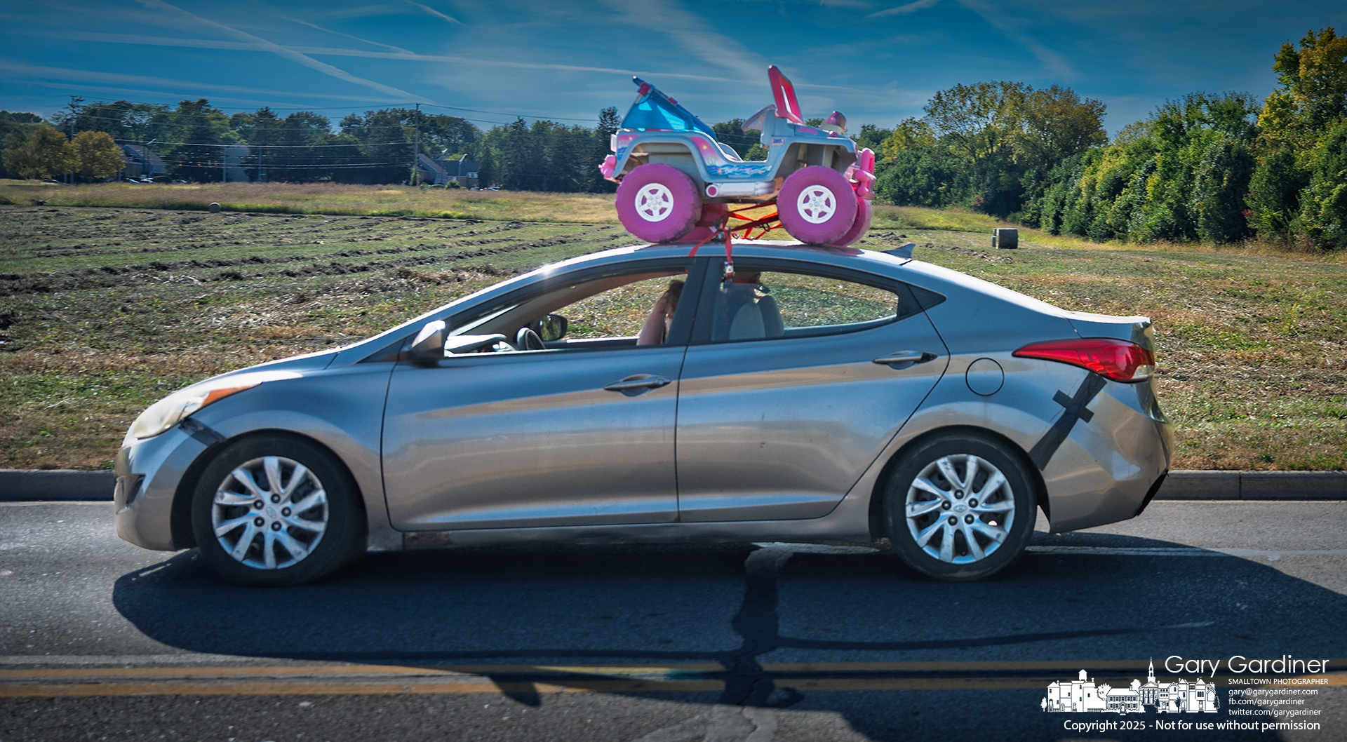 Strapped down with bungee cords and a dose of hope, a pink-and-blue Power Wheel perches atop a sedan cruising down Collegeview Road, past hay fields mid-harvest. My Final Photo for September 9, 2025.