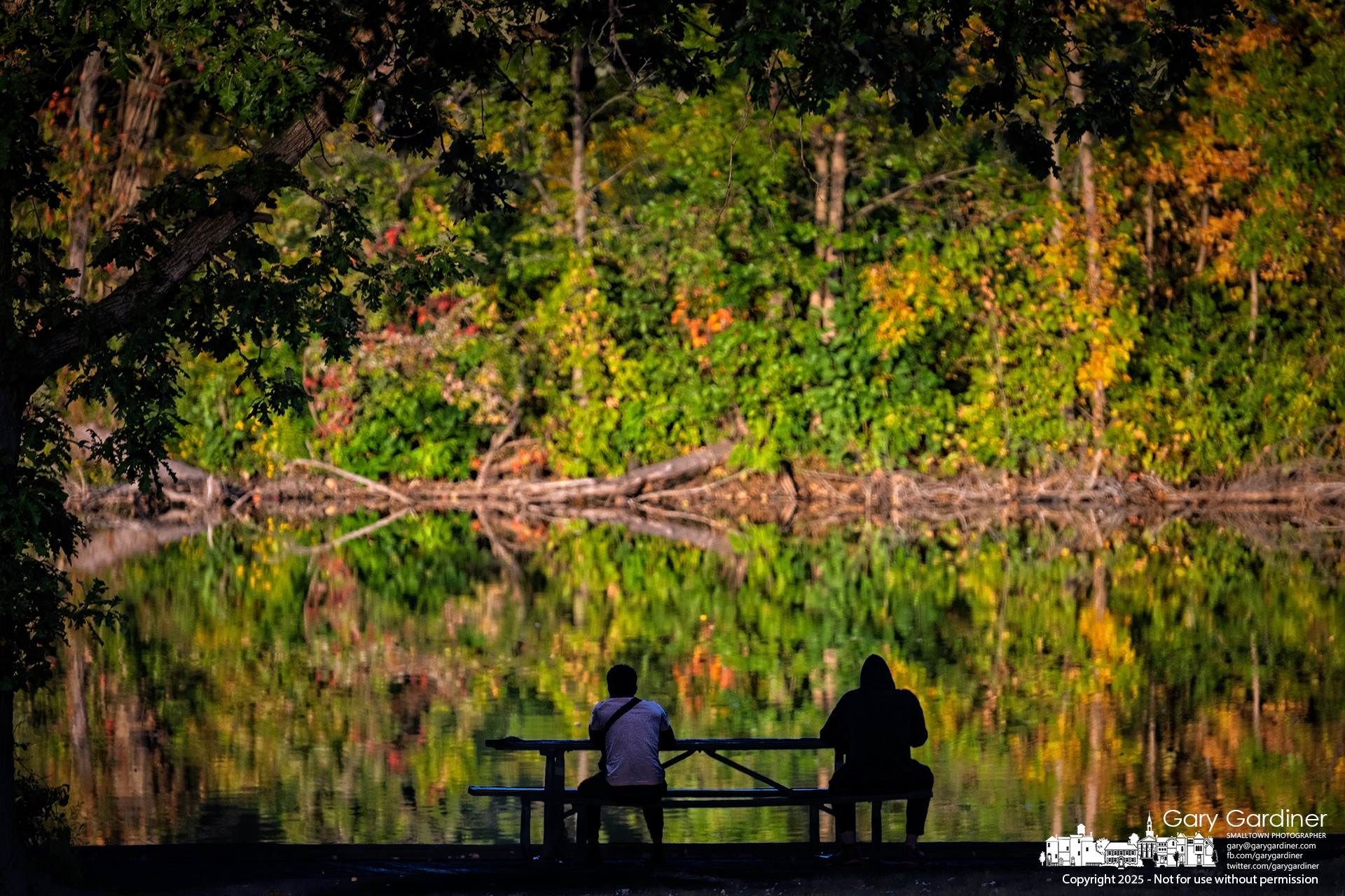 Two people sit quietly at a picnic table in the shade as evening sunlight warms the eastern shore of Schrock Lake at Sharon Woods Metro Park, reflecting the early signs of autumn across the water. My Final Photo for September 18, 2025.