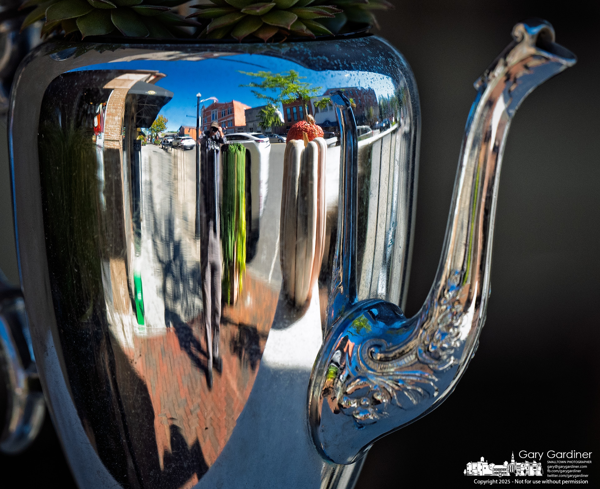 Uptown Westerville is both reflected and distorted in one of the stainless steel pitchers repurposed as planters on the front patio of Asterisk restaurant. My Final Photo for October 21, 2025.