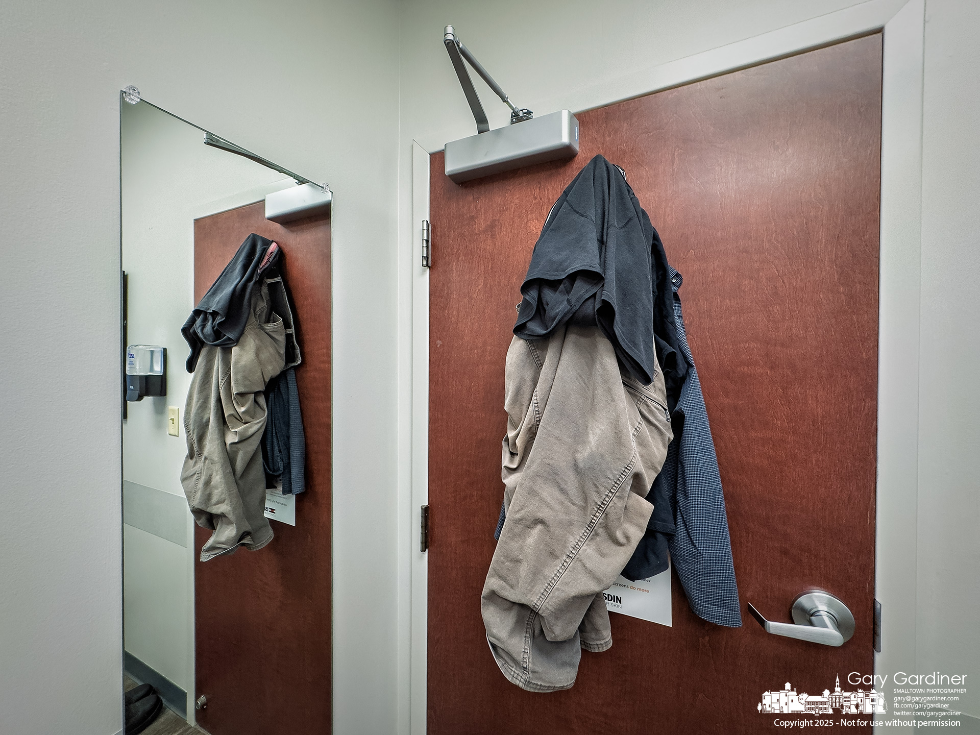 A photographer's clothes hang on hooks on the back of the doctor's examining room door. My Final Photo for October 23, 2025.