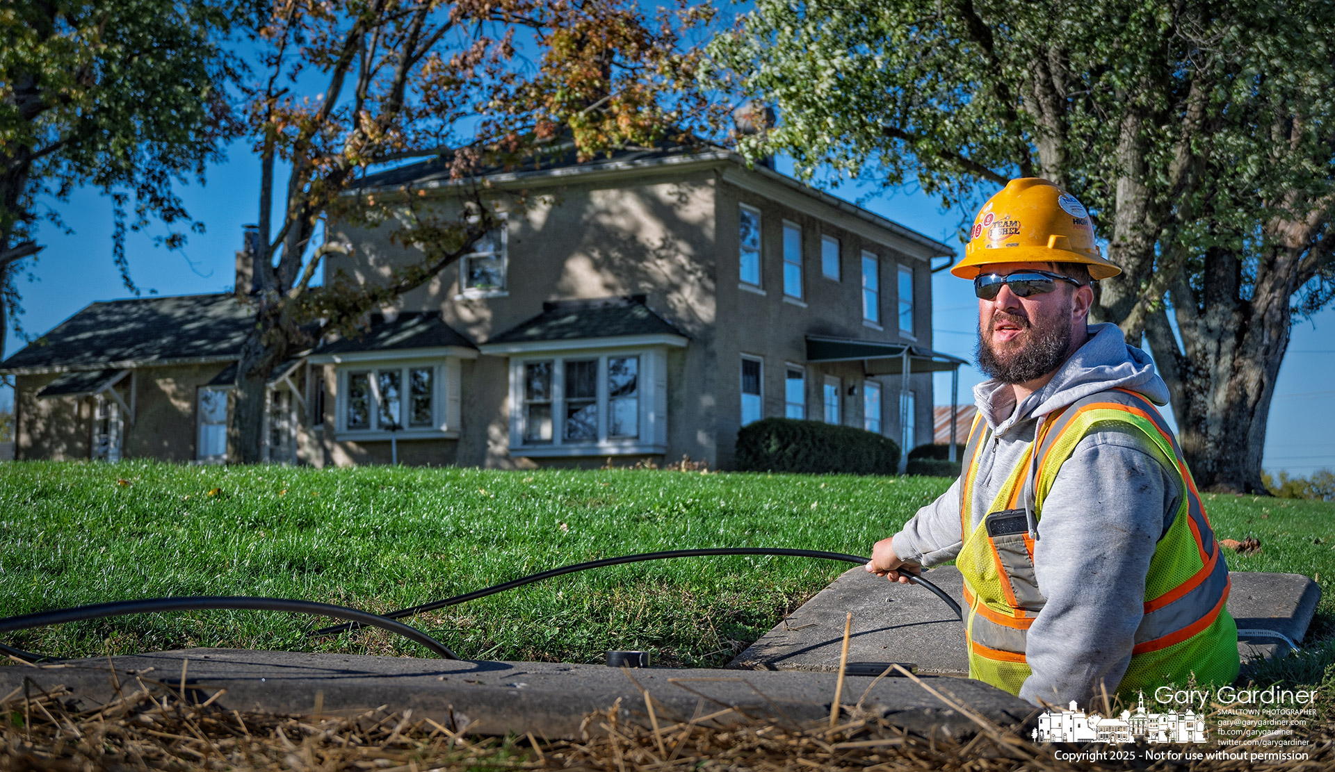 A field technician feeds fiber optic cable into an underground conduit in the right of way in front of the historic Yarnell Farm house. My Final Photo for October 20, 2025.