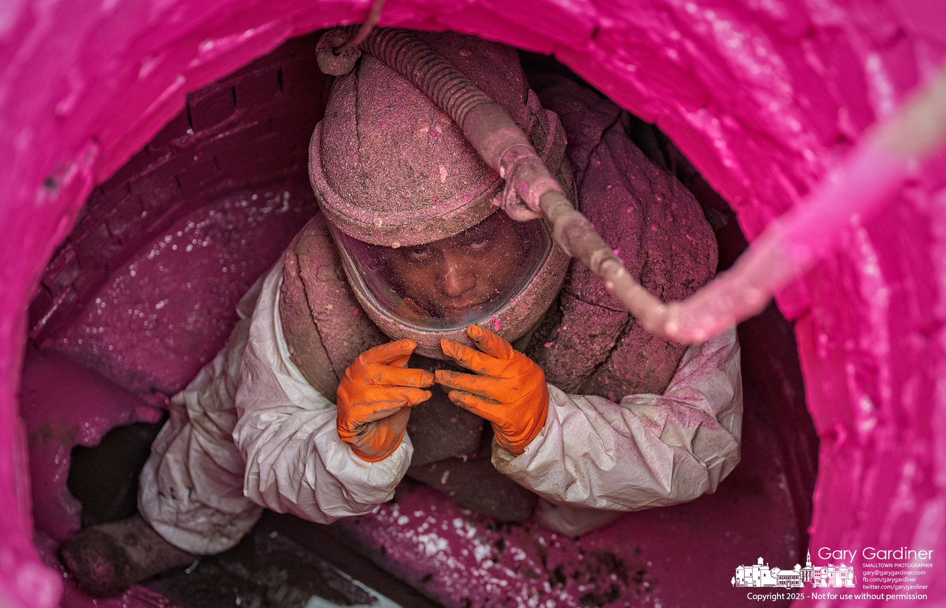 A worker, coated in pink spray adhesive and white foam residue, looks up from inside a storm sewer in Uptown. Wearing a positive air pressure hood for protection, he applies a PVC liner to aging brick manholes as part of a city infrastructure project to extend their lifespan. My Final Photo for October 2, 2025.