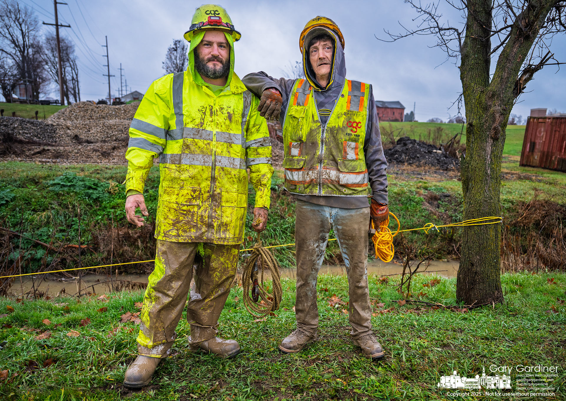 Two muddy workers pose for a photo at the end of a rainy Tuesday, after making no progress installing a concrete culvert because the creek remained flooded. My Final Photo for November 25, 2025.